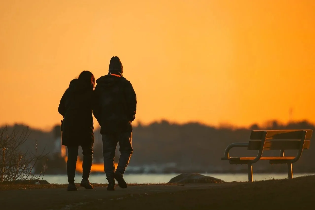 Two people walking together at sunset near water, with a bench on the right.