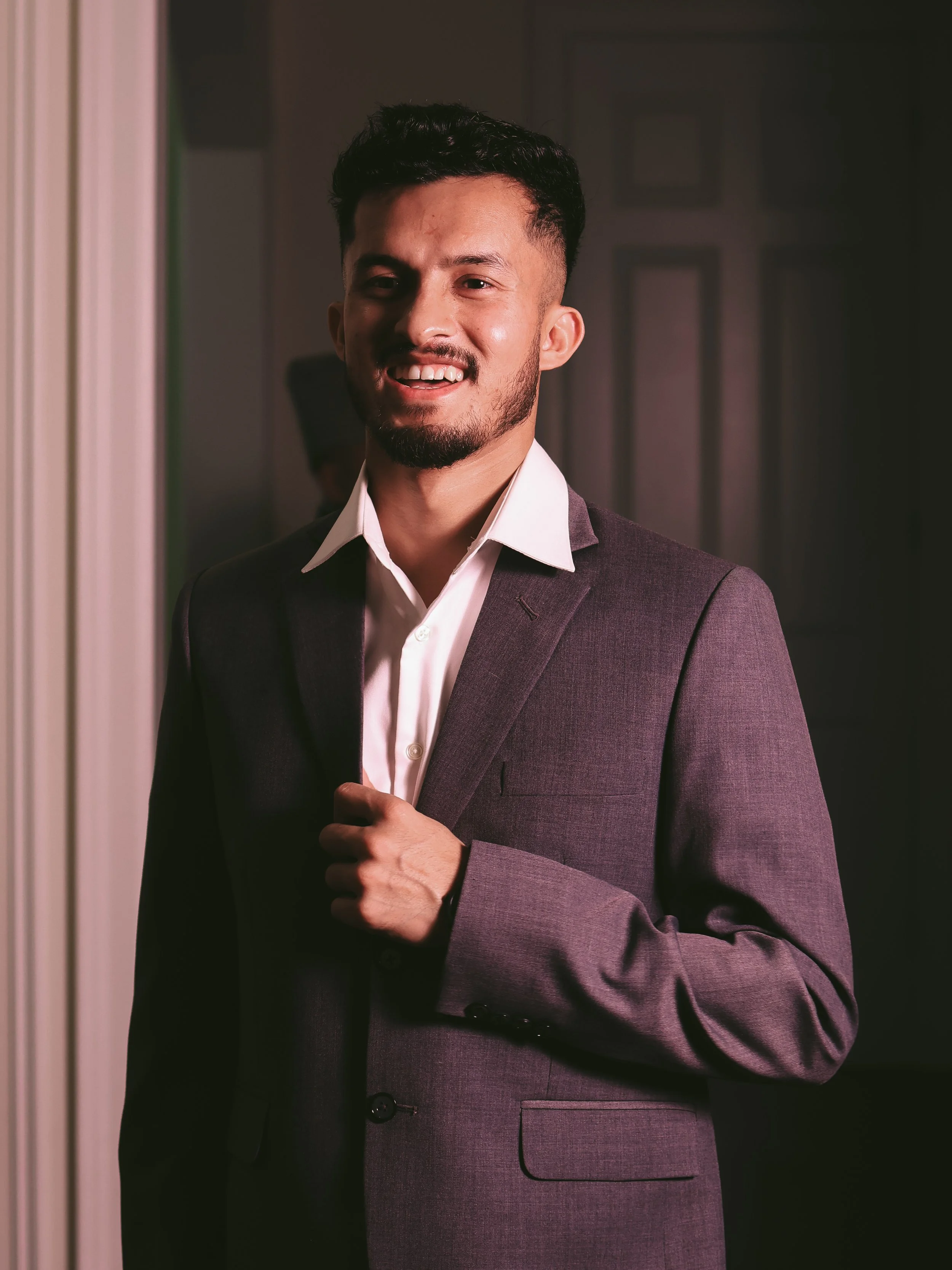 A man wearing a gray suit with a white shirt, smiling and adjusting his jacket in a dimly lit interior setting.