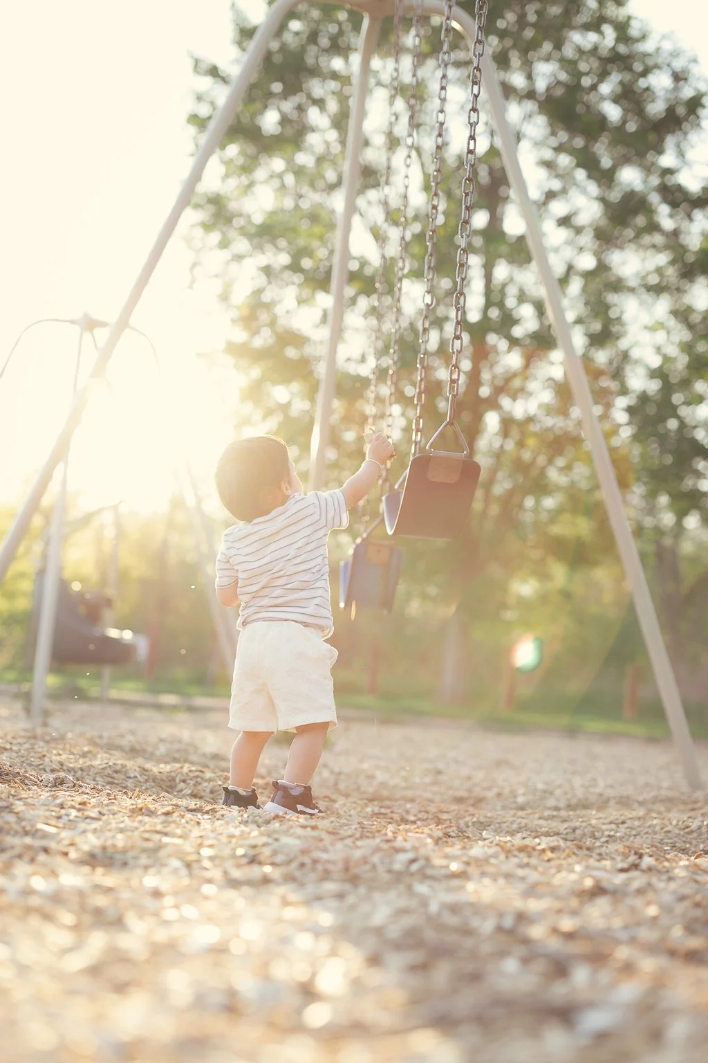 A young child in a striped shirt and shorts playing on a swing set in a park during sunset.