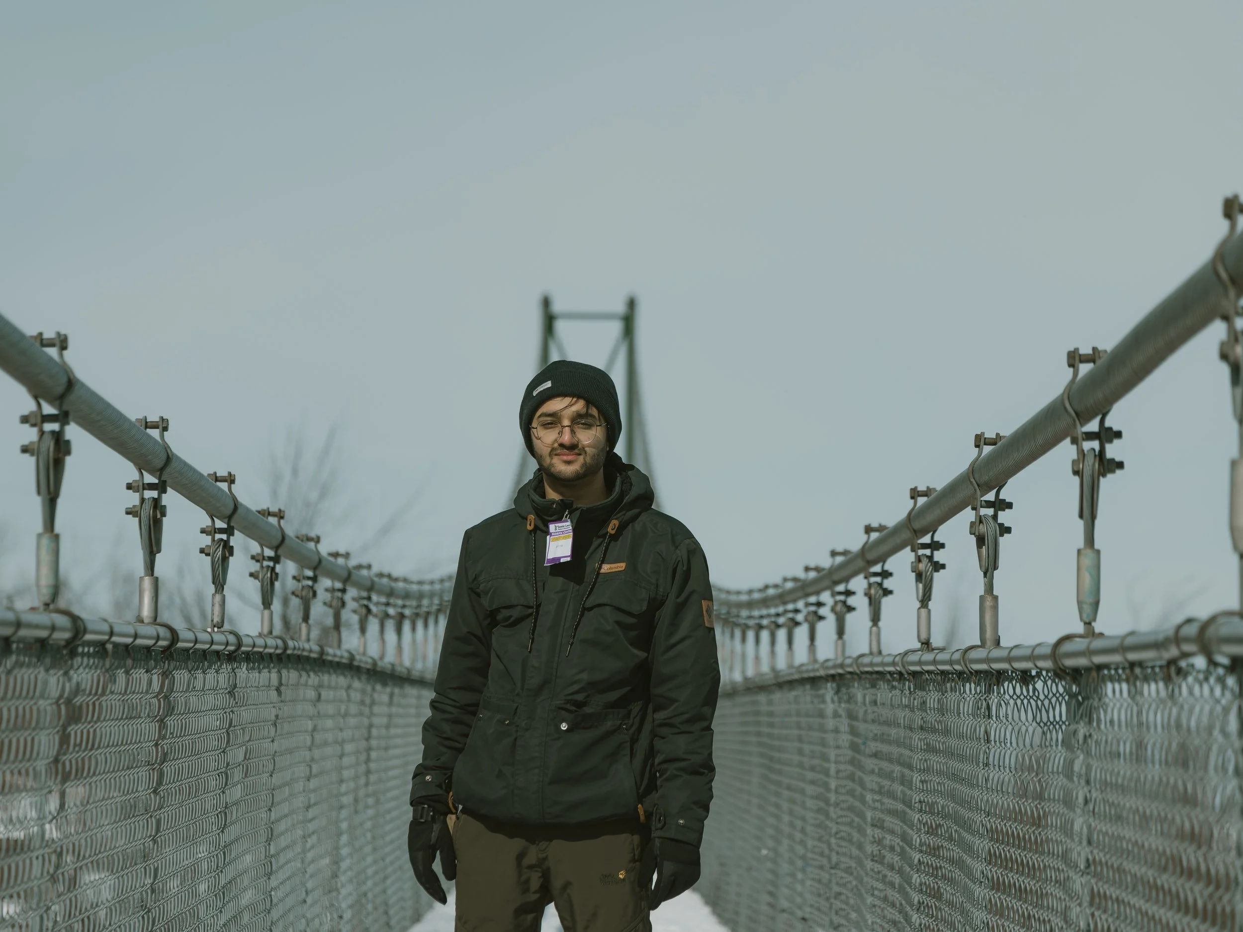Young man wearing glasses, a black beanie, and a black jacket standing on a suspension bridge with metal railings, overcast sky in the background.