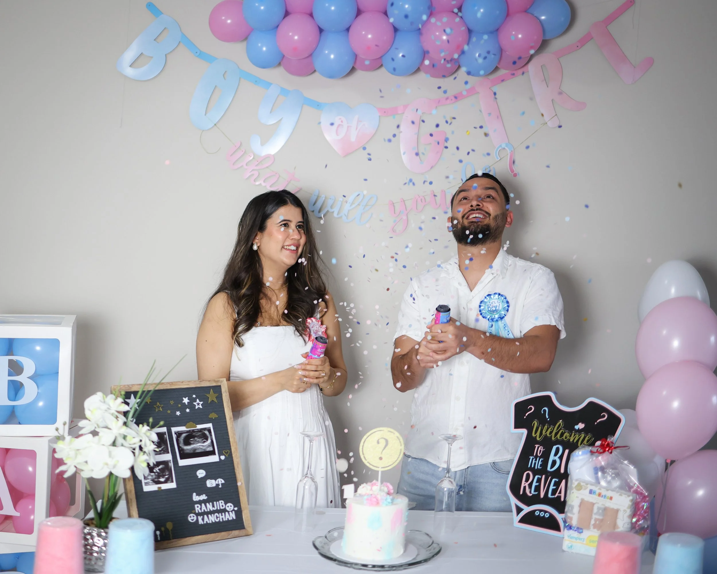 A baby shower celebration with a woman and a man smiling, surrounded by pink and blue balloons, balloons, confetti, and decor including a welcome sign, memorabilia, and ultrasound pictures.