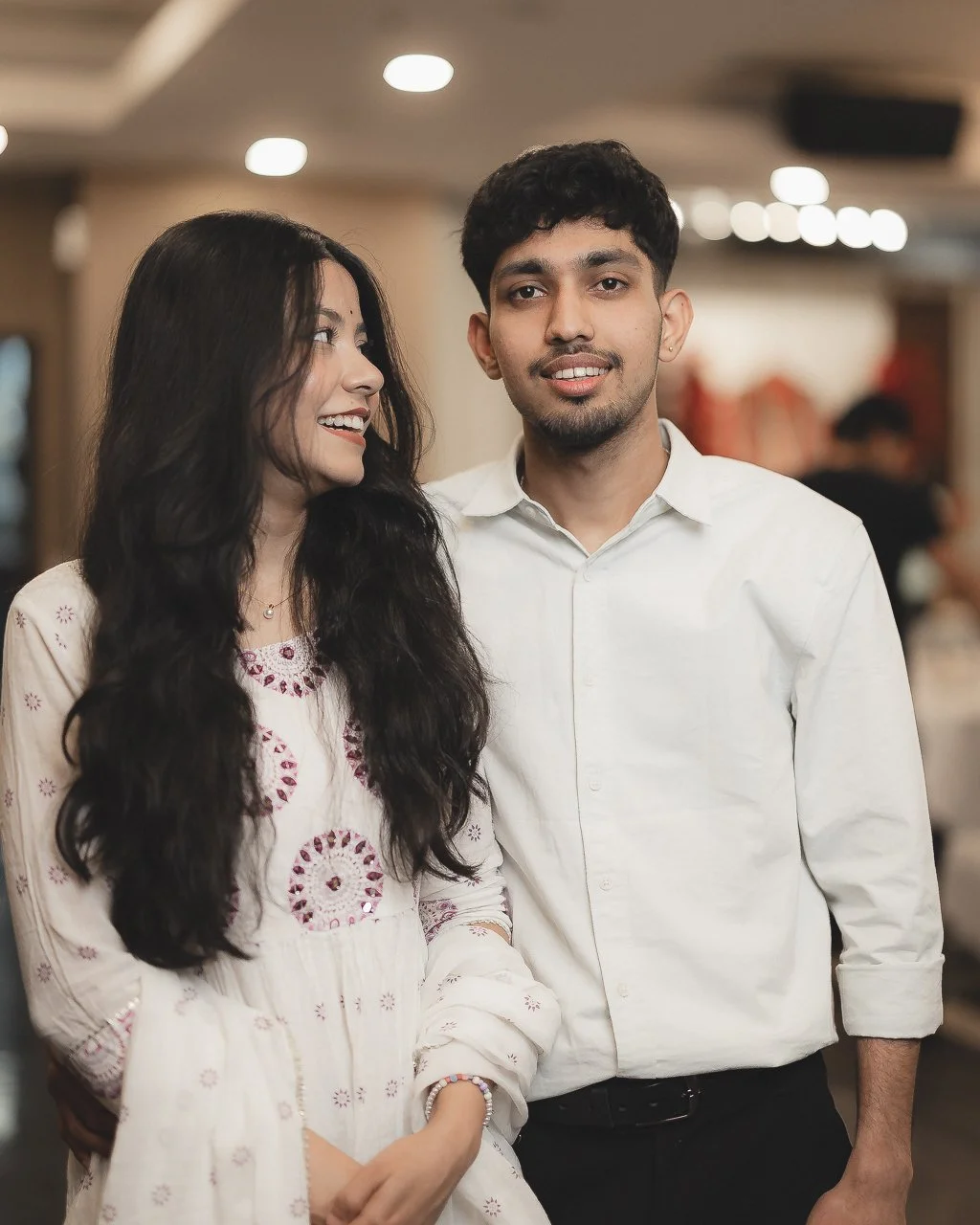 A young man and woman standing together indoors, smiling and looking at each other, casually dressed in white shirts, with blurred background.