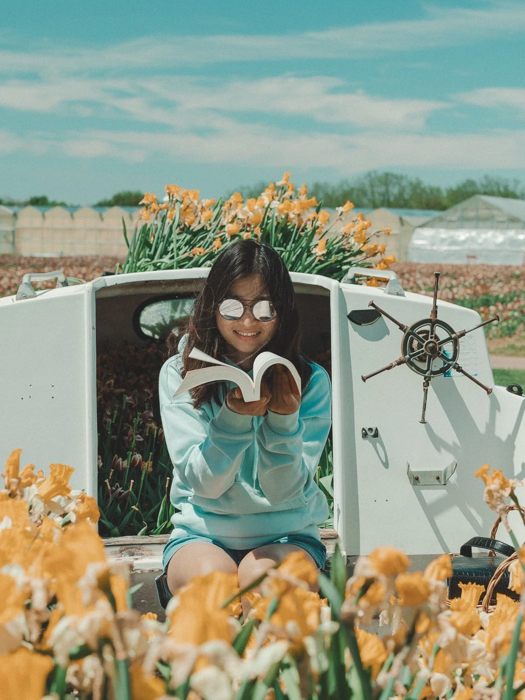A girl sitting in front of a white cart decorated with yellow flowers, reading a book, with sunglasses on, outdoors on a sunny day with a blue sky.
