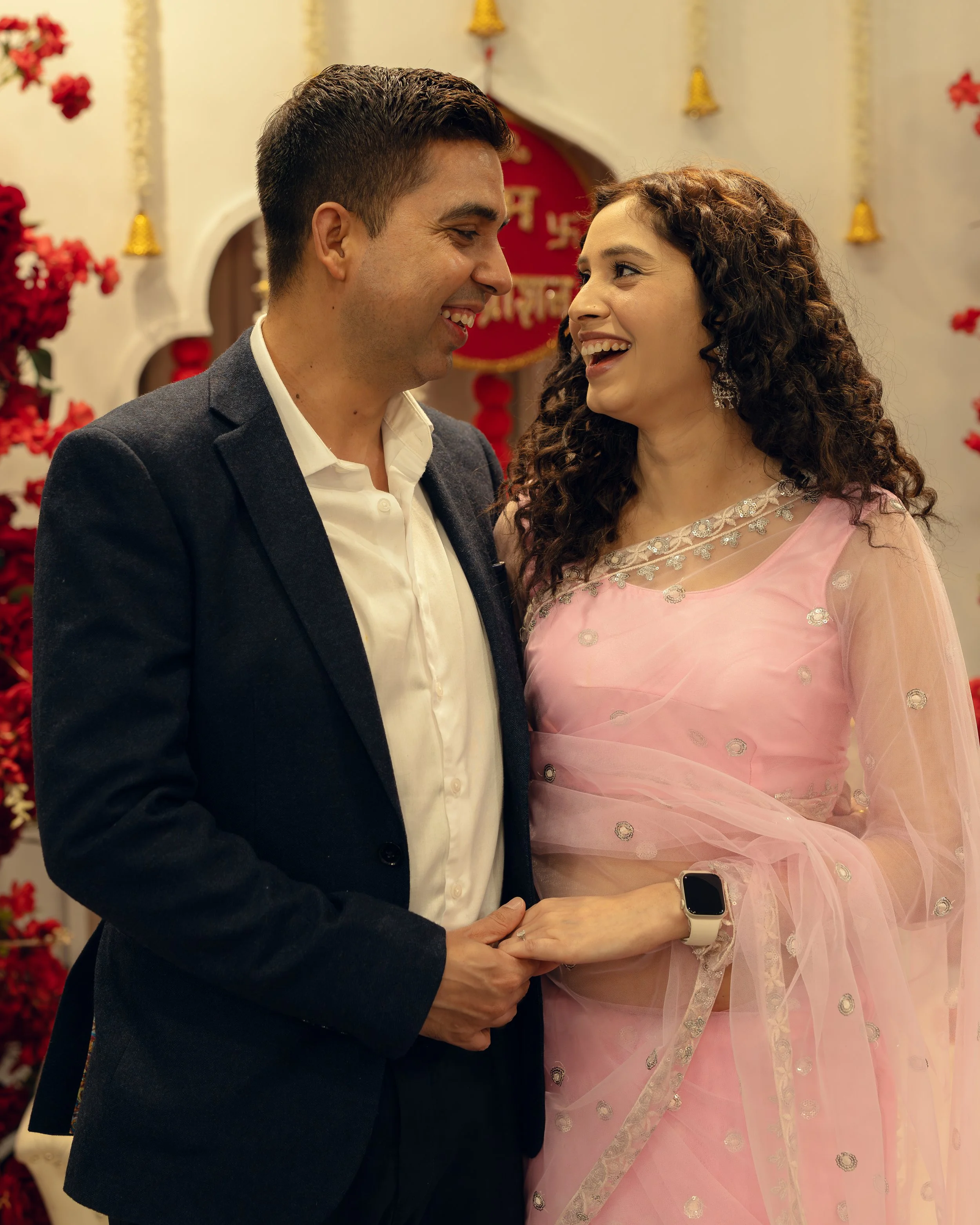 A man and woman share a joyful moment, standing close and smiling at each other, surrounded by festive decorations like red flowers and small bells.