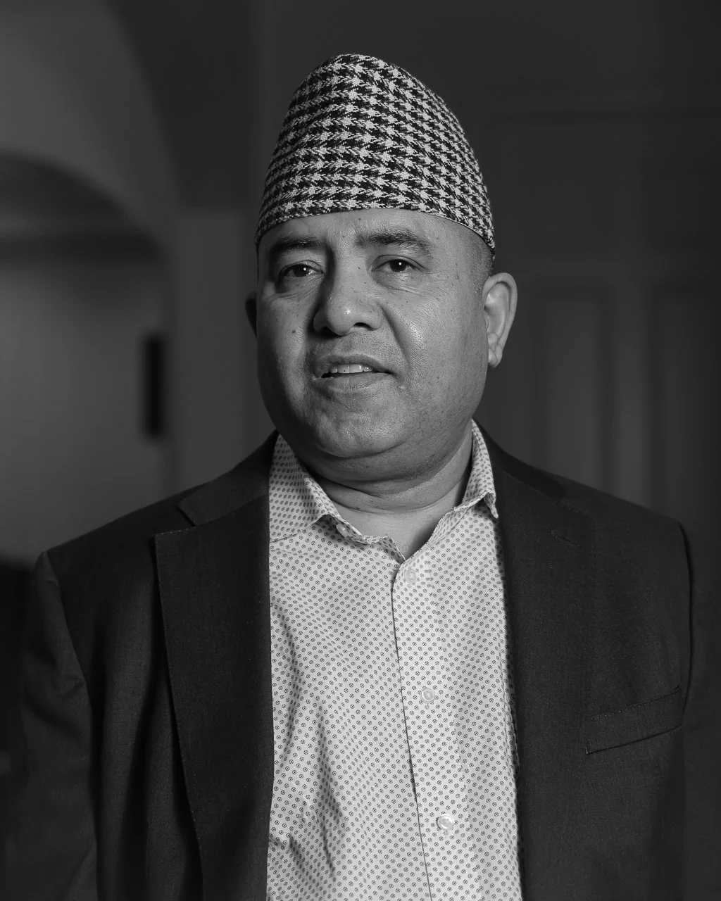 A man wearing a patterned hat, a suit jacket, and a patterned shirt, speaking or listening attentively in an indoor setting.
