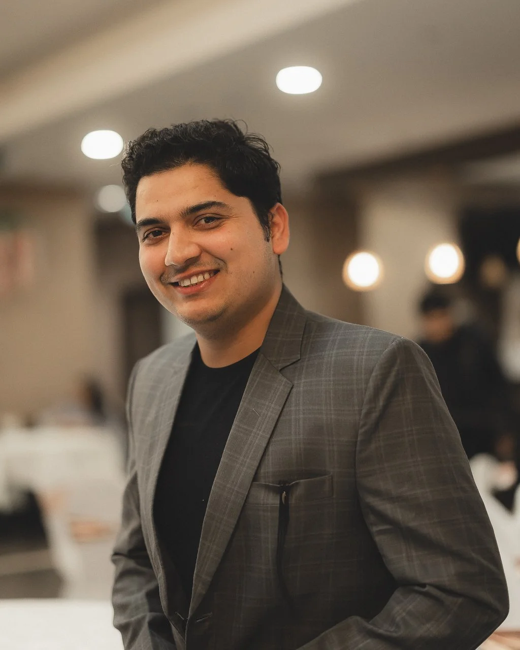 A smiling man with dark hair in a suit jacket standing in a warmly lit indoor setting.