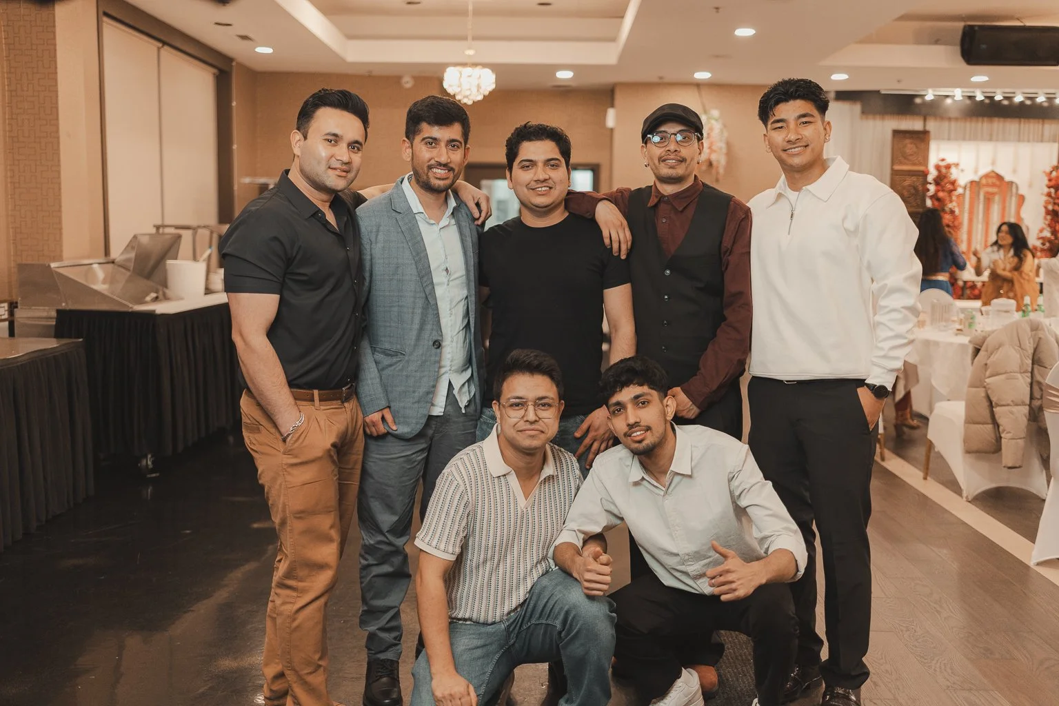 A group of nine young men posing together in a banquet hall, with five standing in the back and two kneeling in front, smiling at the camera.