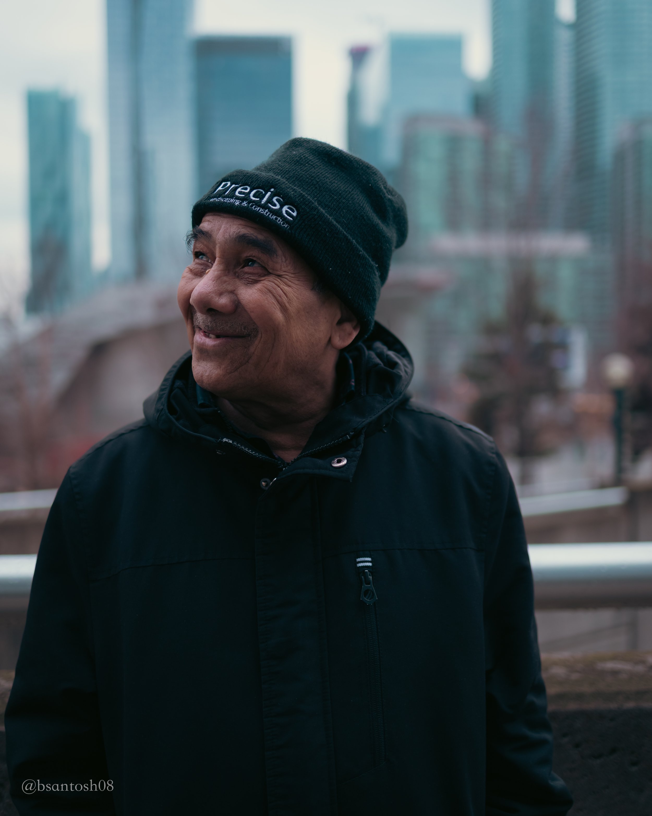Close-up of an elderly man smiling, wearing a black beanie and jacket, standing outdoors with blurred city buildings in the background.