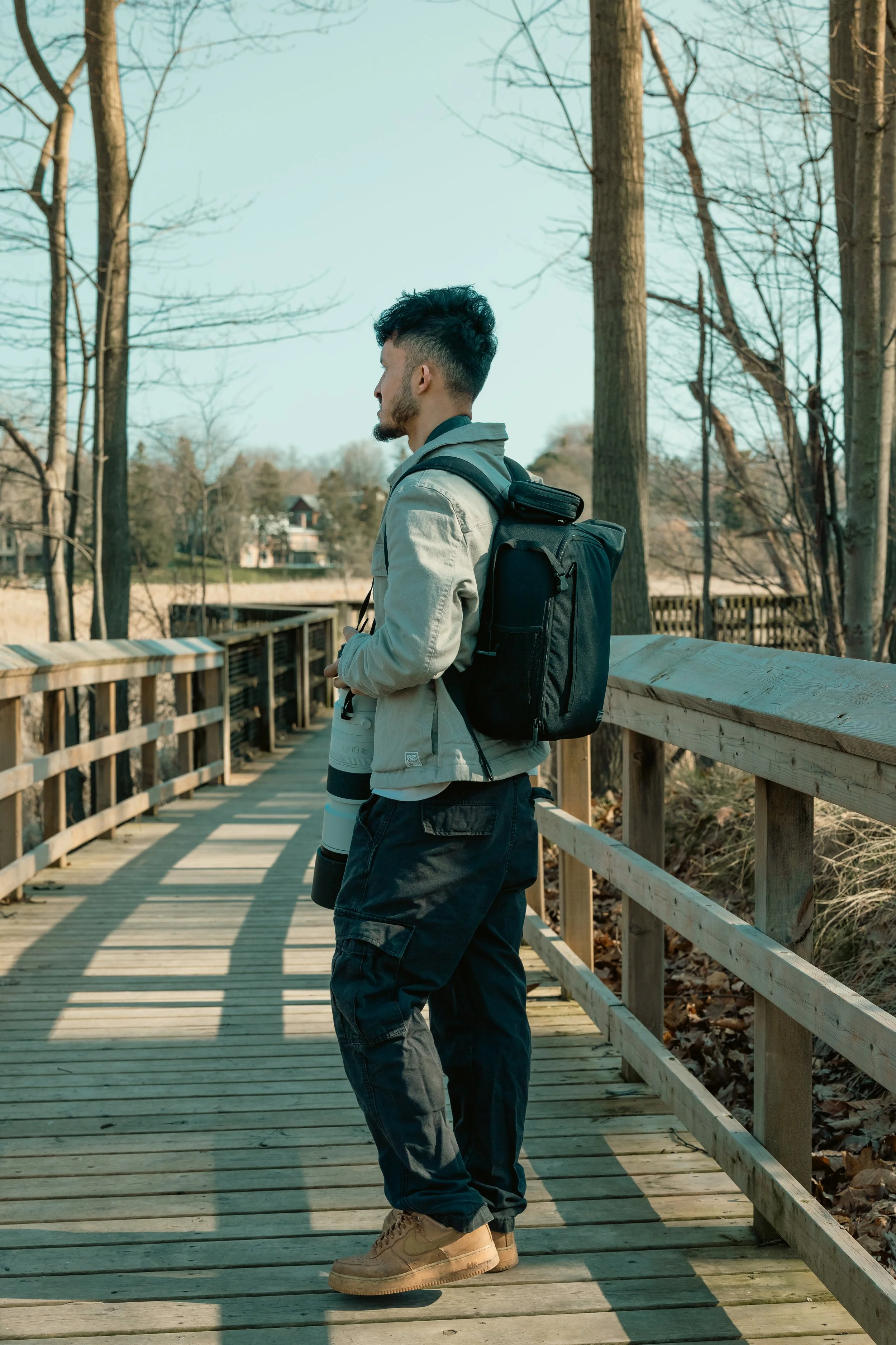 Young man with a camouflage jacket and black backpack standing on a wooden bridge, holding a camera lens, surrounded by bare trees during daytime.