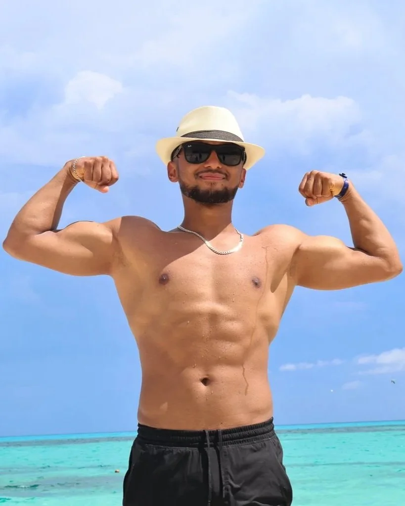 A shirtless man wearing sunglasses, a white hat, and a necklace, flexing his biceps on a beach with clear turquoise water and blue sky in the background.