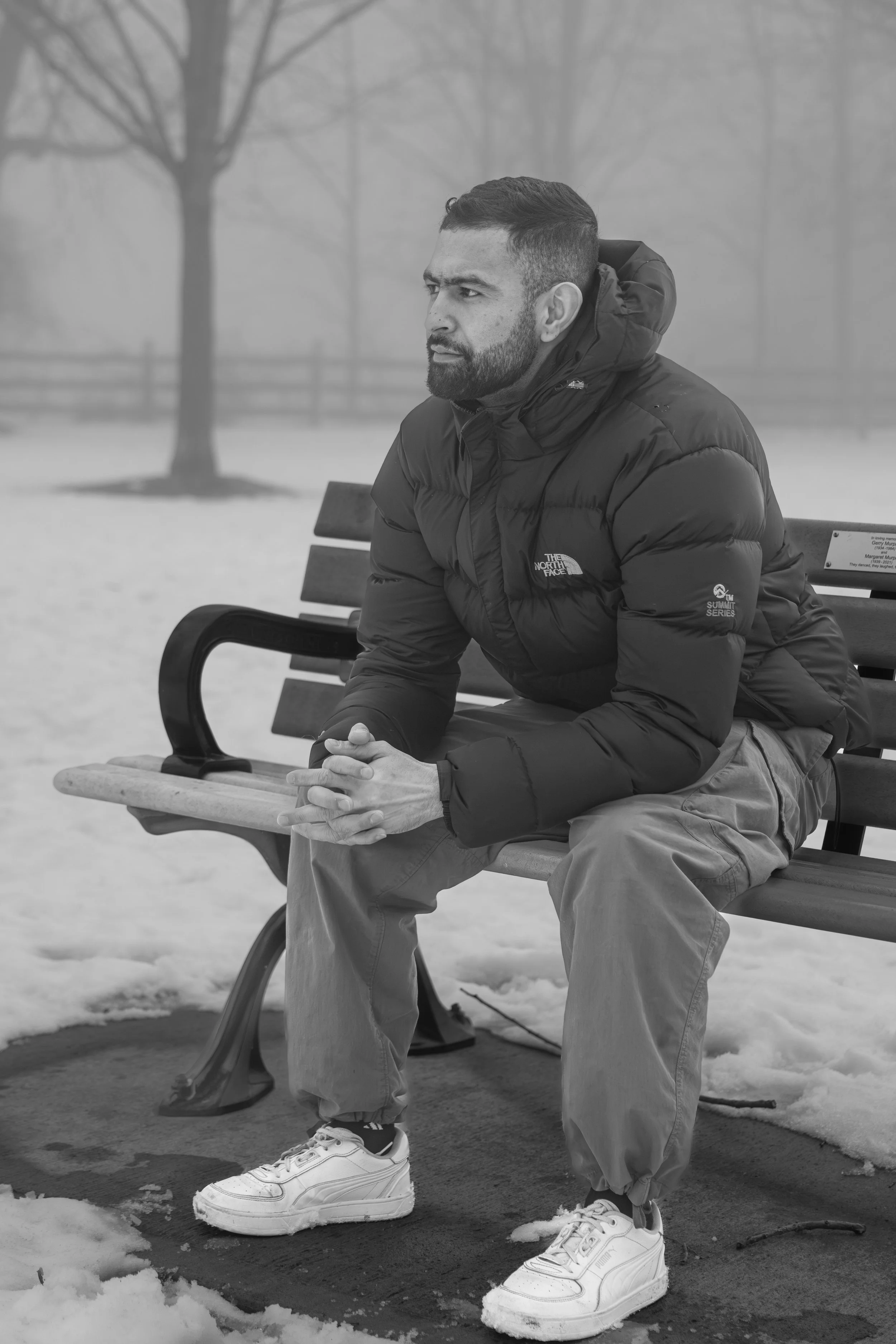 A man sitting on a park bench outdoors on a cloudy, foggy winter day, wearing a puffy jacket and sneakers, with snow on the ground and bare trees in the background.