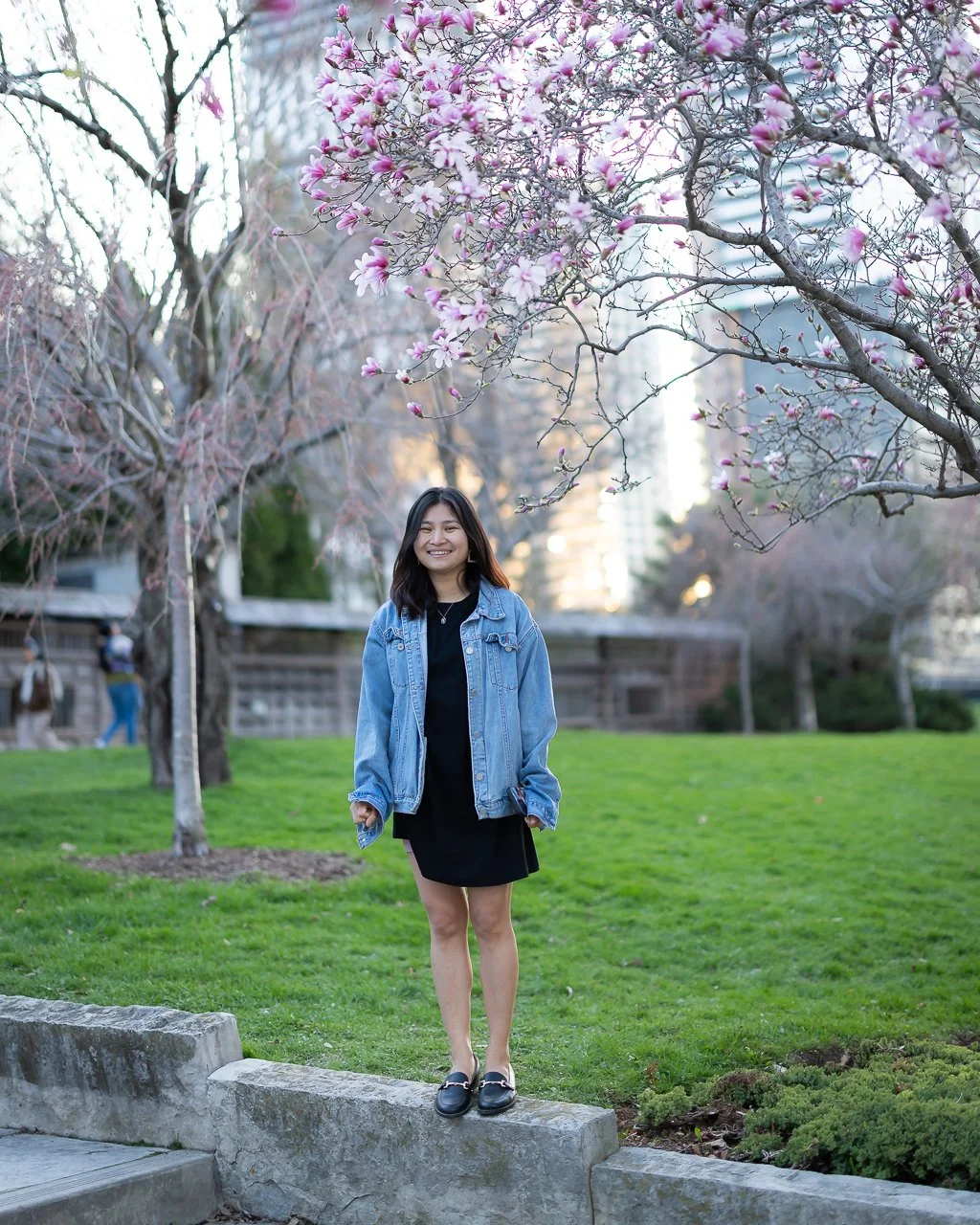 A young woman smiling and standing on a concrete ledge in a park during springtime, with pink magnolia blossoms on trees behind her.
