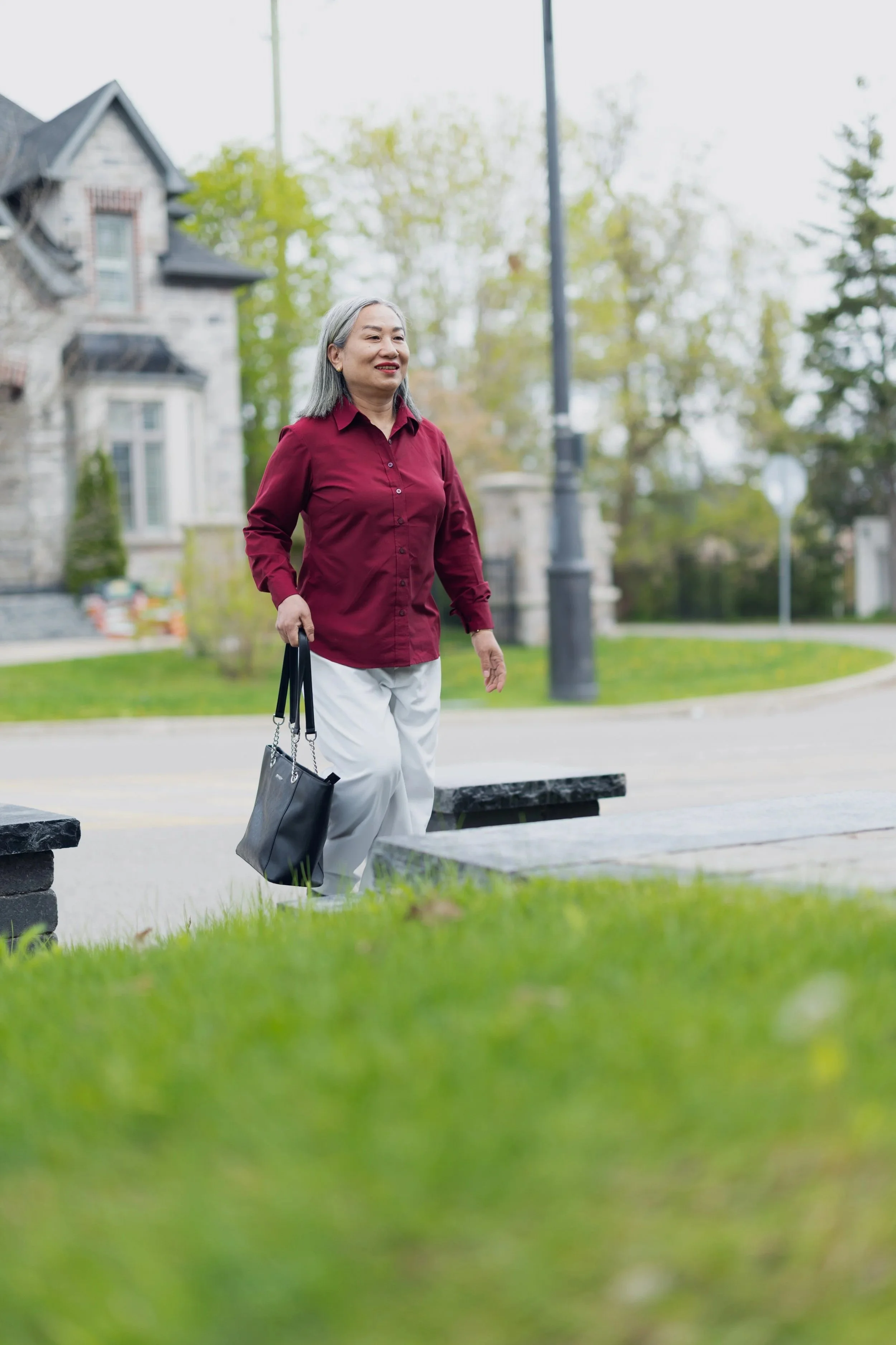 A woman with gray hair wearing a red blouse and white pants walking in a park and carrying a black handbag.