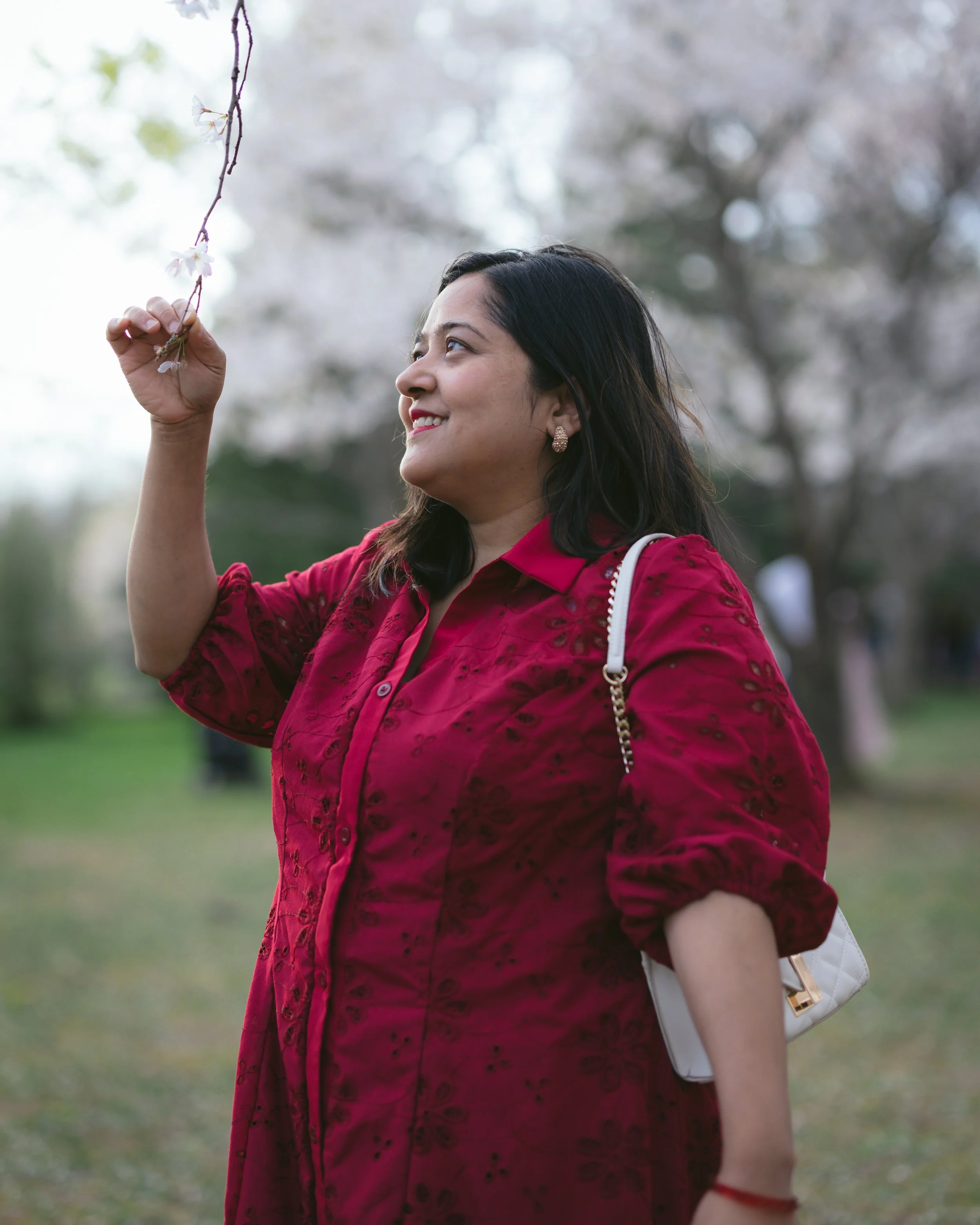 A woman wearing a red kurta with embroidery details, holding a small white purse with a chain strap, standing outdoors in front of blooming trees, smiling and looking up at a flowering branch.