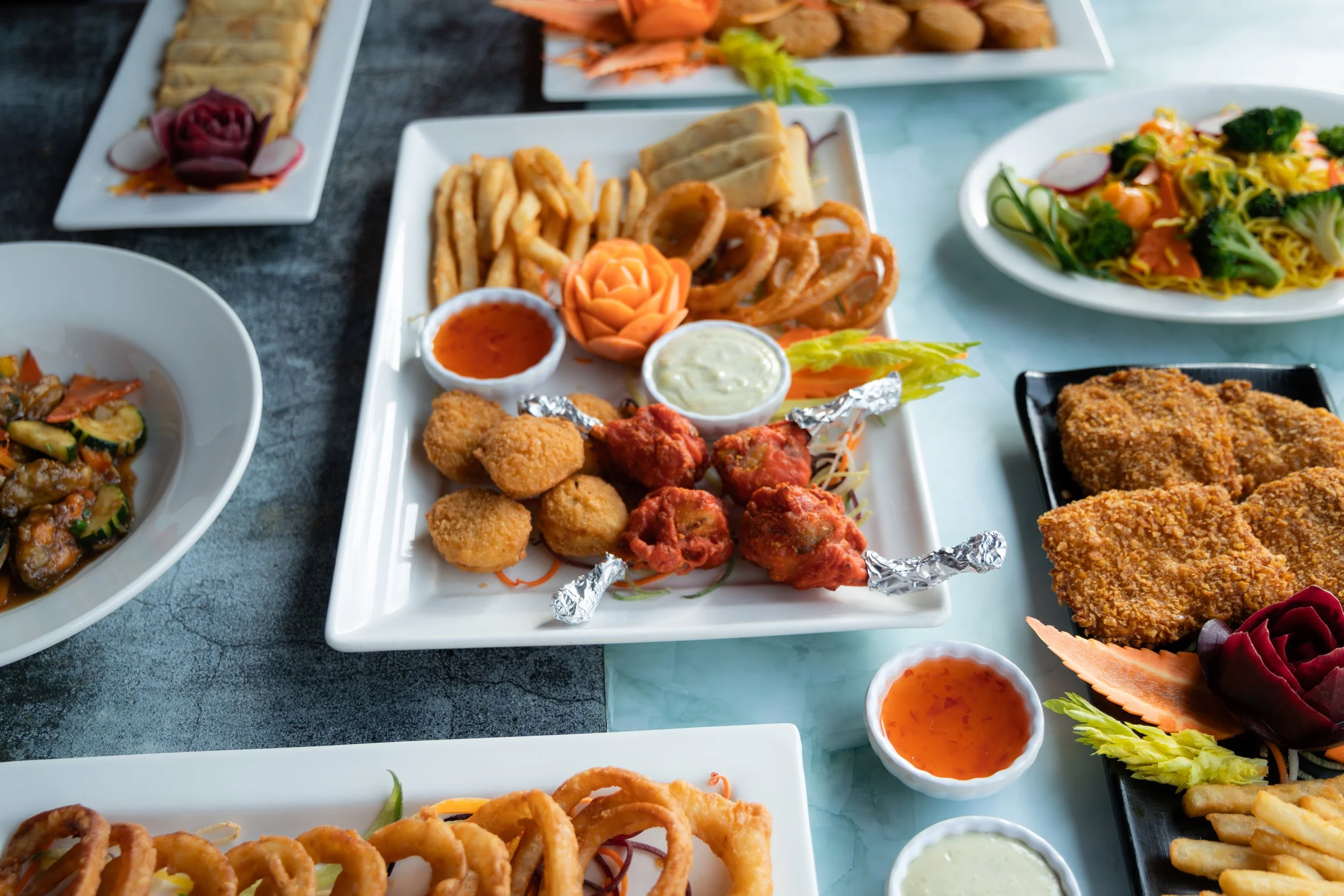 Assorted dishes including fried foods, vegetables, and dipping sauces on white and black plates, with some garnishes such as carved vegetables, set on a dark and a teal surface.