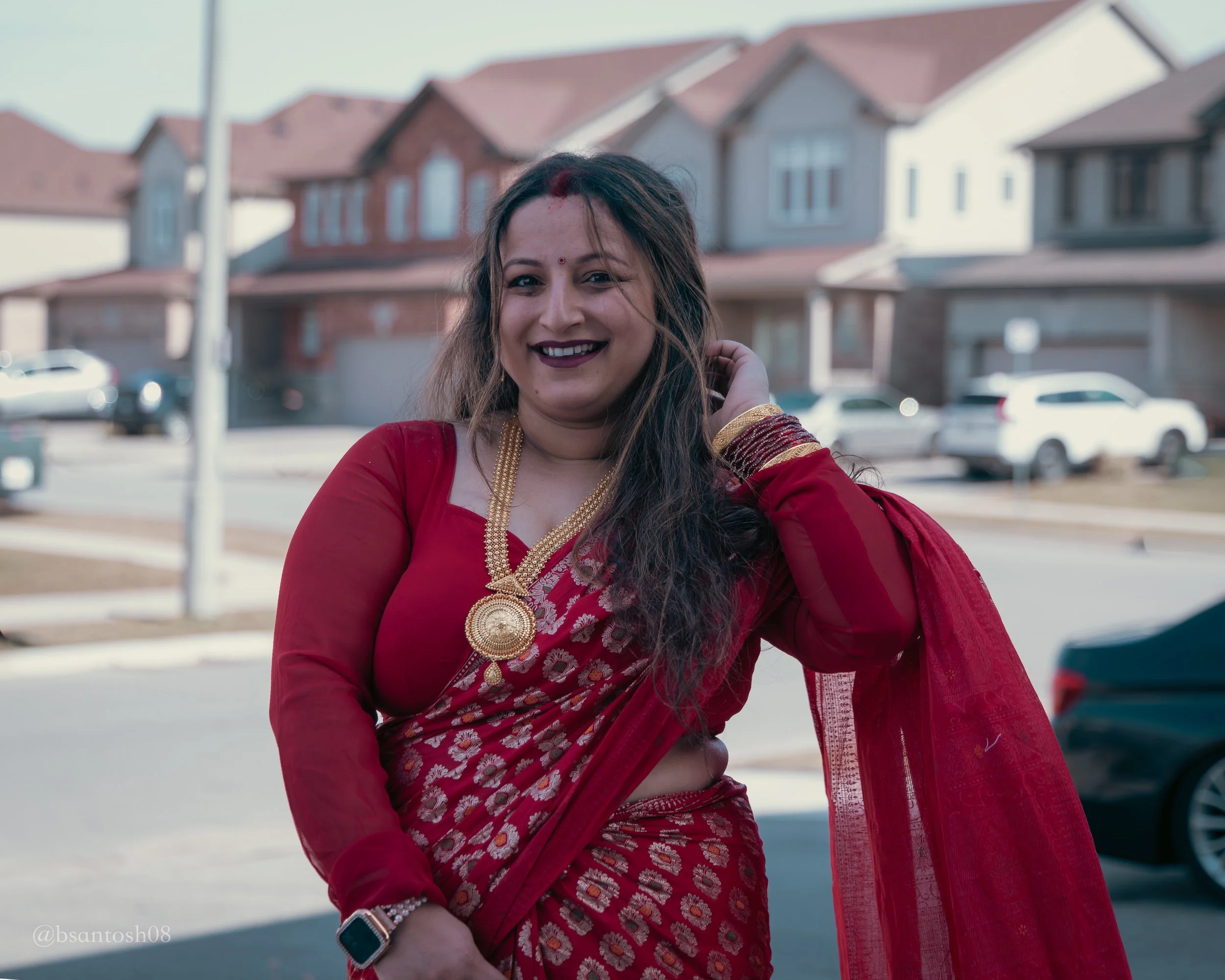 A woman dressed in a red traditional Indian outfit, smiling outdoors in front of residential buildings and parked cars.