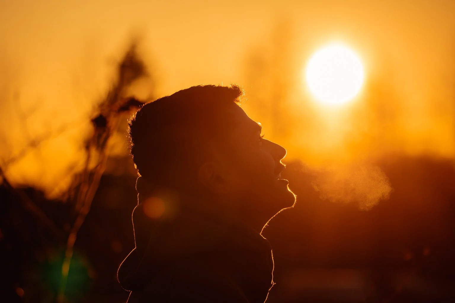Silhouette of a person in profile blowing out mist or breath against a setting sun with an orange sky.