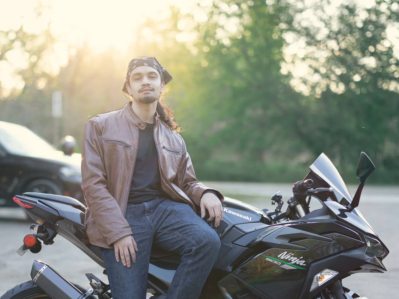 A man sitting on a black Kawasaki Ninja 650 motorcycle outdoors with greenery and trees in the background, and the sun shining through the leaves.