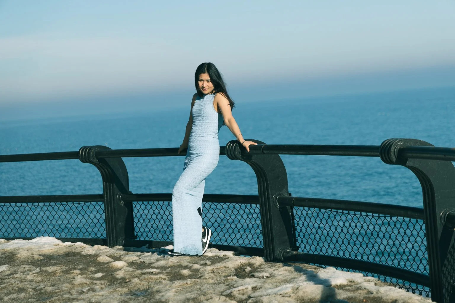 A woman in a light blue outfit standing by a black railing on a rocky surface near the ocean, with a clear blue sky in the background.
