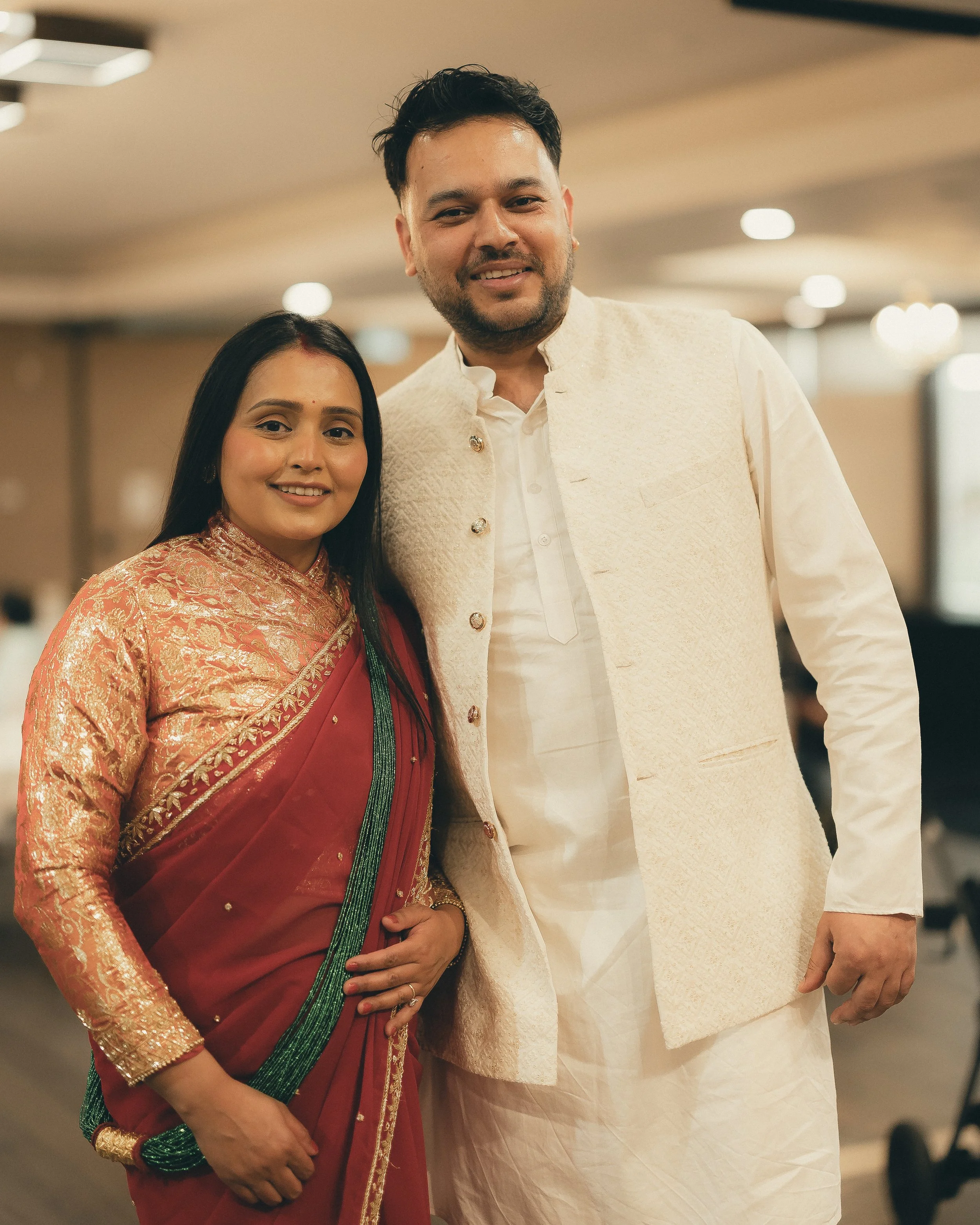 A man and woman dressed in traditional Indian attire, smiling and posing indoors with blurred lighting and background.