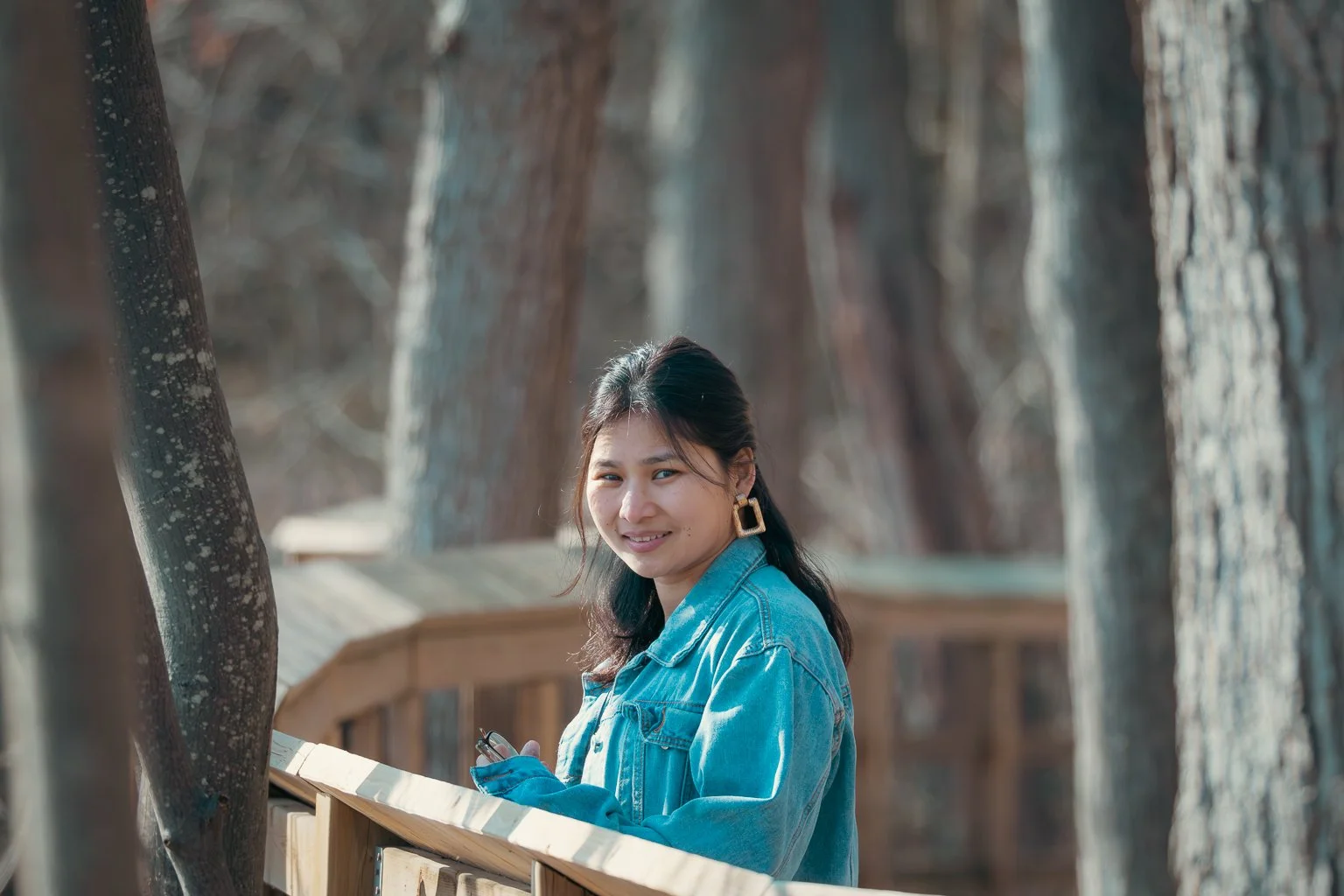 A young woman with dark hair in a ponytail, wearing a denim jacket and gold earrings, standing outdoors on a wooden bridge, holding a smartphone and smiling at the camera with a background of tree trunks.