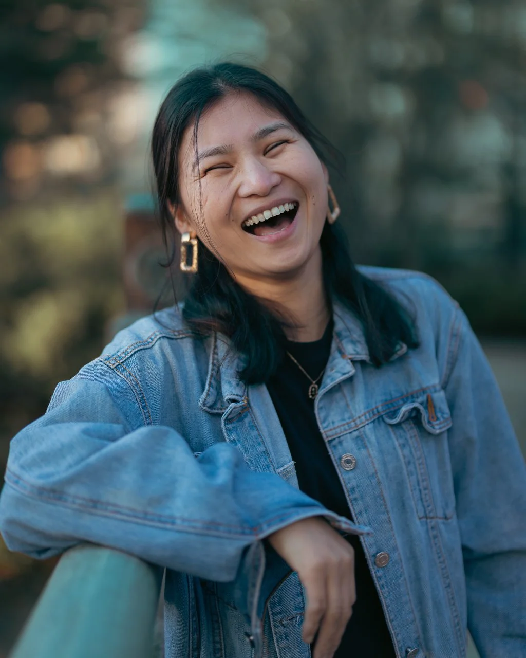 A woman with medium-length black hair, wearing earrings and a denim jacket, is smiling and laughing outdoors.