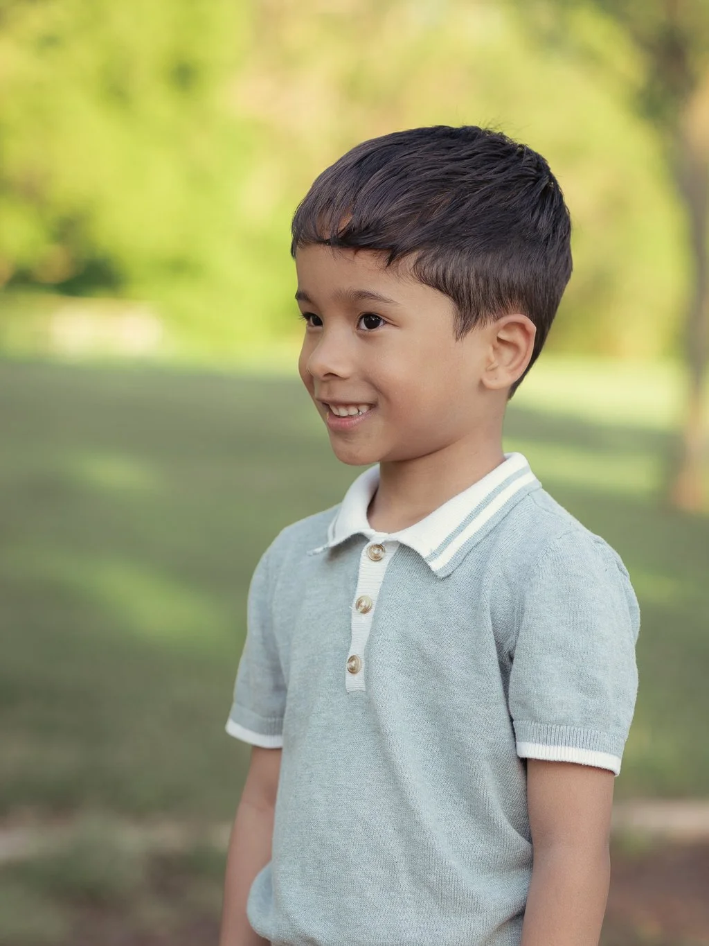 A young boy with dark hair smiling outdoors in a park with blurred green trees and grass in the background.