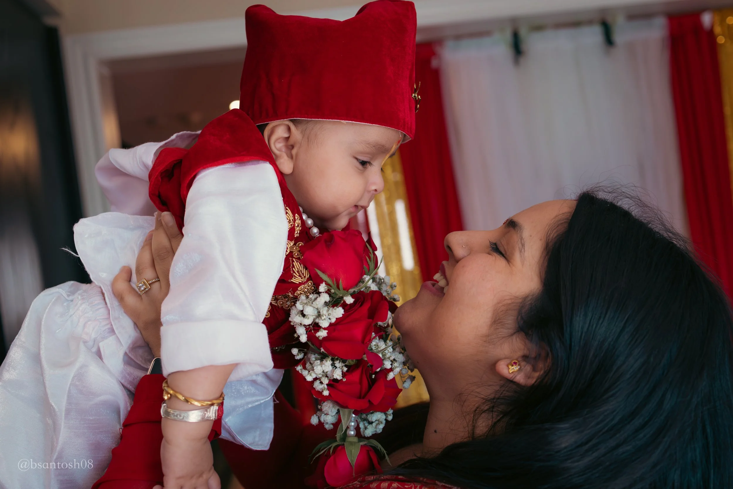 A young child dressed in traditional red and white attire is being held up by a woman with dark hair. They are touching foreheads and smiling, holding a bouquet of red and white flowers. The background features curtains with red, white, and gold colors.