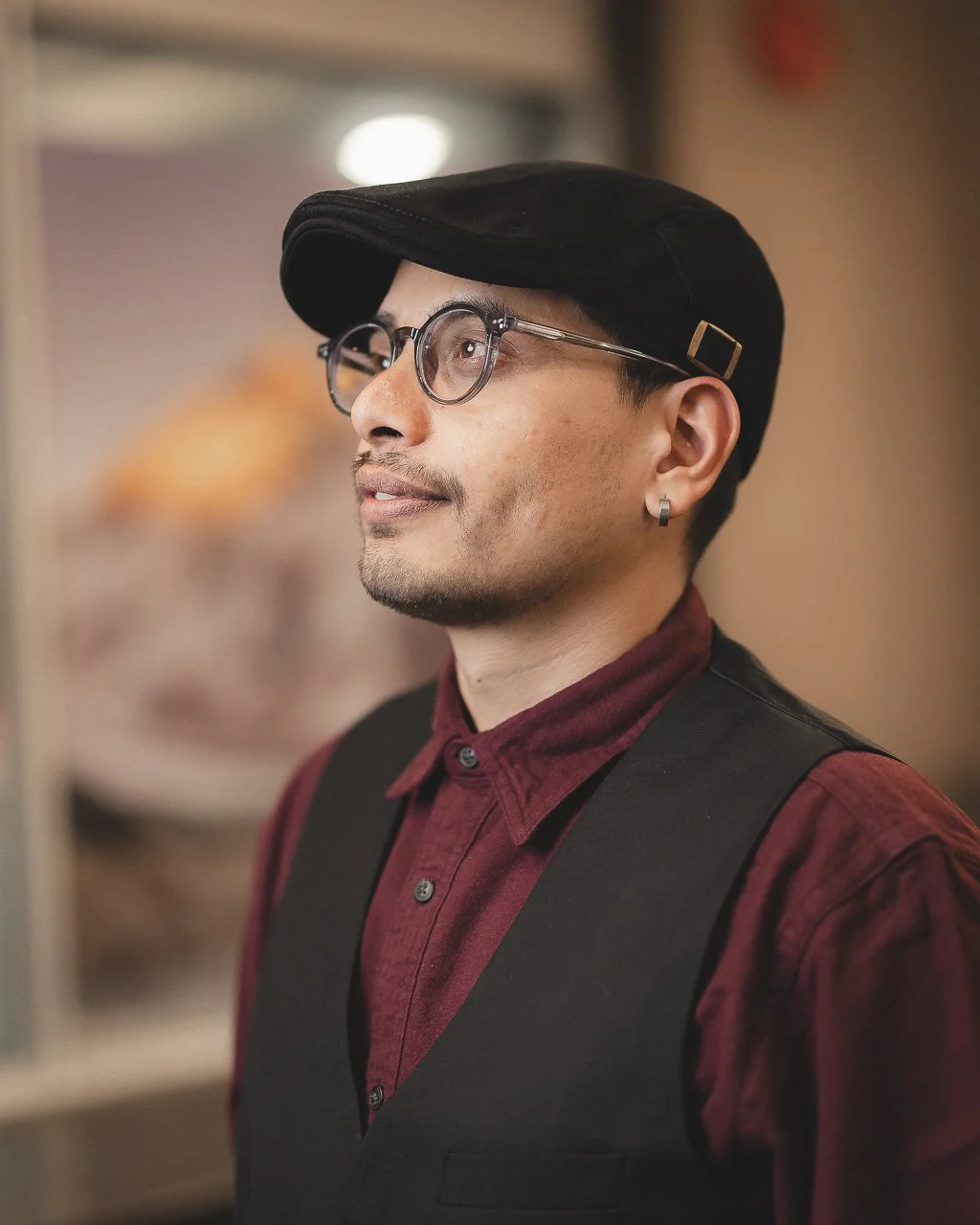 A young man wearing glasses, a black cap, a maroon shirt, and a black vest, looking slightly to the side indoors.