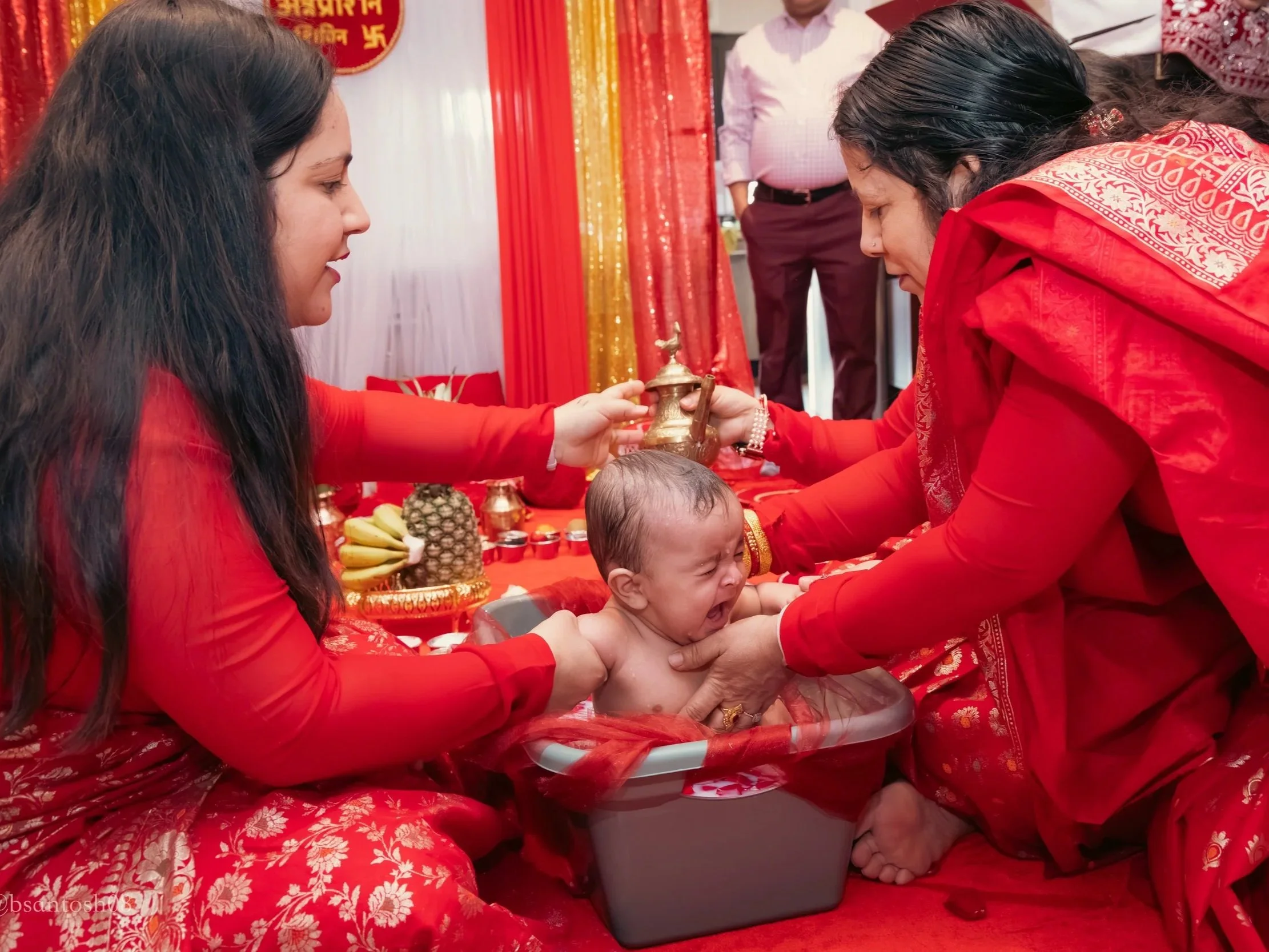A woman is performing a Hindu ritual called baby naming ceremony on a baby in a metal tub, with another woman holding an oil lamp over the baby, in a decorative room with red and gold drapes.