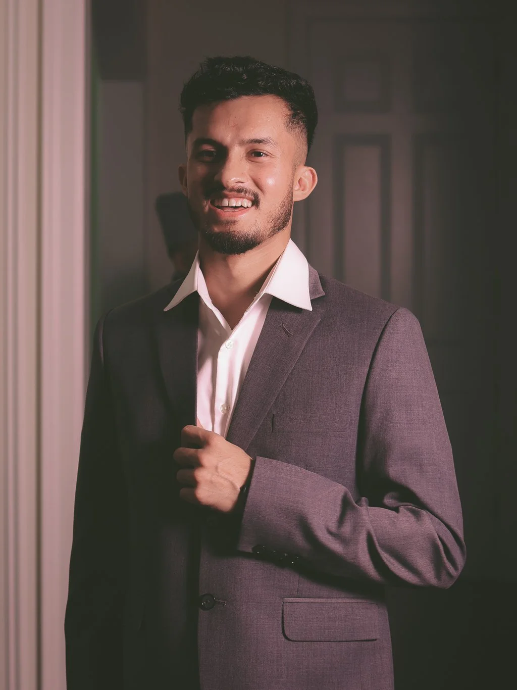 A man in a dark suit and white shirt smiling indoors.
