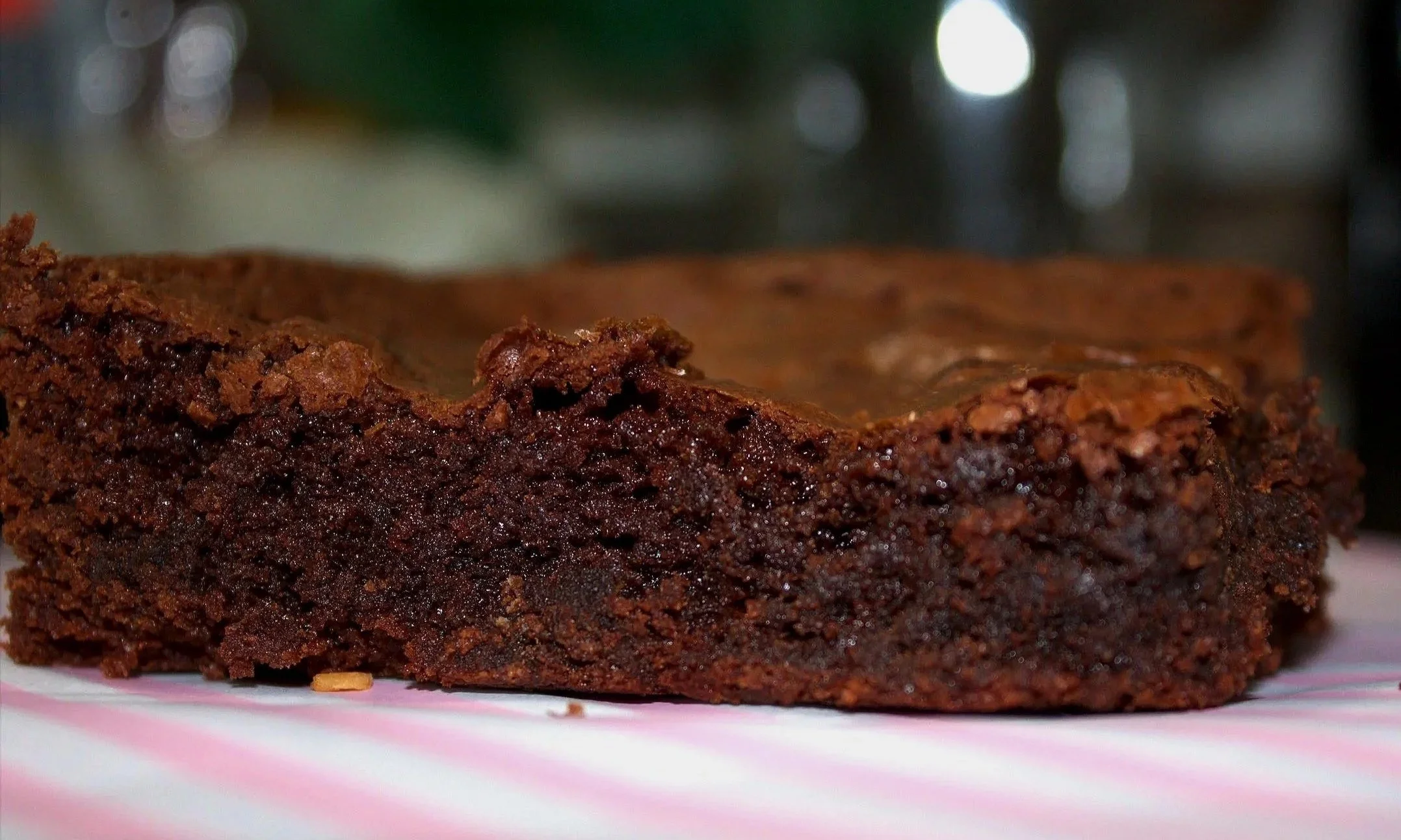 Close-up of a chocolate brownie on a white and pink plate with a blurred dark background and bright lights.