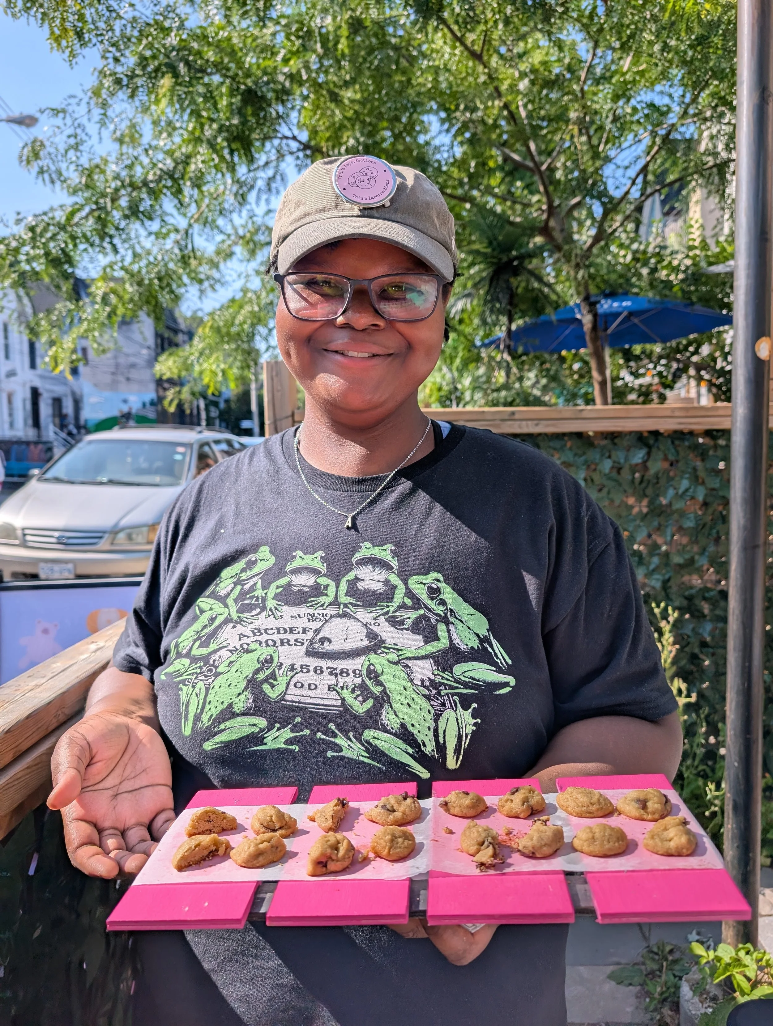 Owner and founder, Trinity with glasses and a green cap, smiling and holding a pink tray of cookies outdoors on a sunny day, with green trees and a blue umbrella in the background.