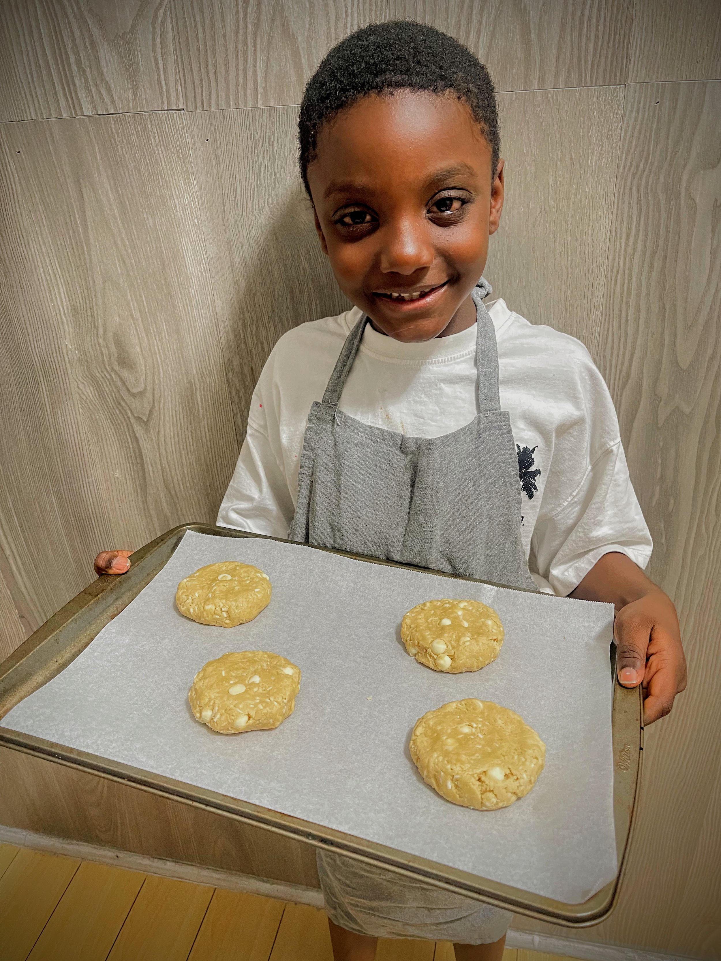 A young boy wearing a white shirt, gray apron, and khaki shorts holding a metal baking tray with 4 unbaked cookies on parchment paper. The cookies are round and slightly uneven.