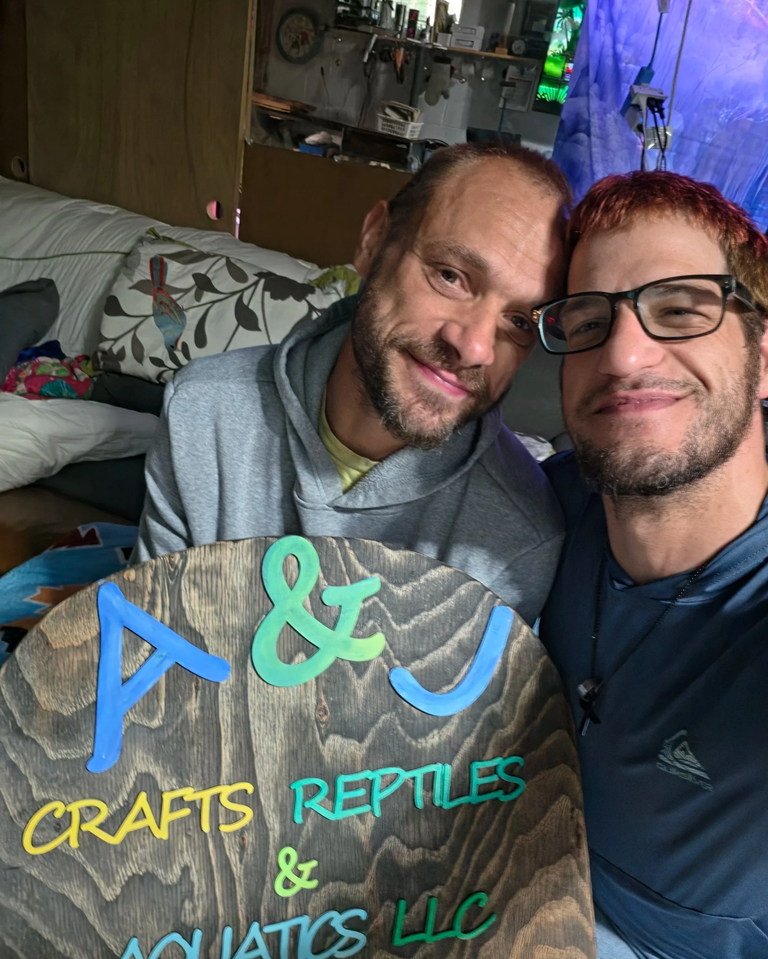 Two men smiling and taking a photo together, with a decorated wooden sign in the foreground that says 'Reptiles and Amphibians' in colorful letters, in a room with a blue and purple backdrop.