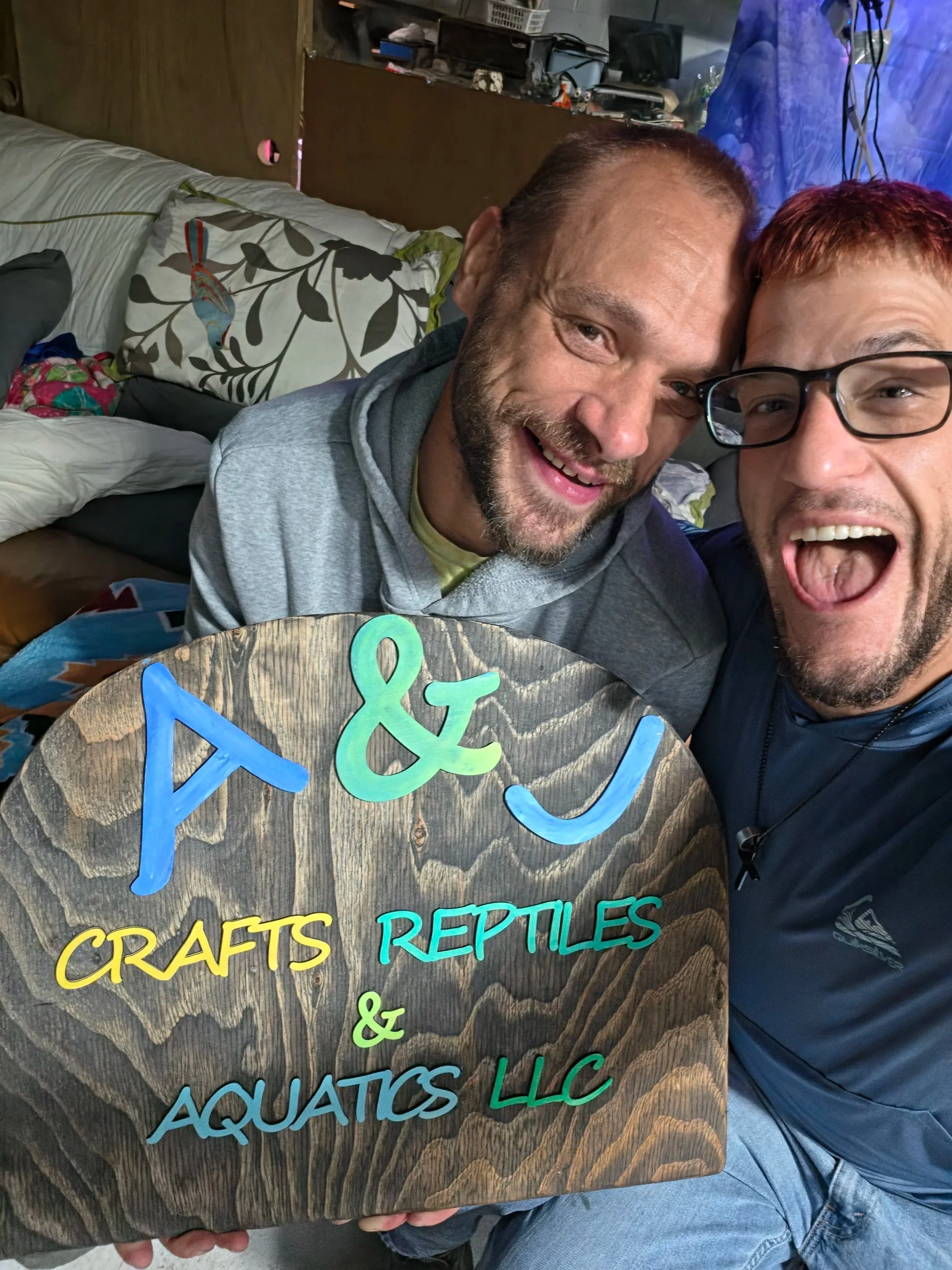 Two men with happy expressions posing in front of a wooden sign that reads 'Crafts & Reptiles'. One man has a beard and is wearing a gray hoodie, the other is wearing glasses and has reddish hair. The background appears to be a cluttered room with various items.