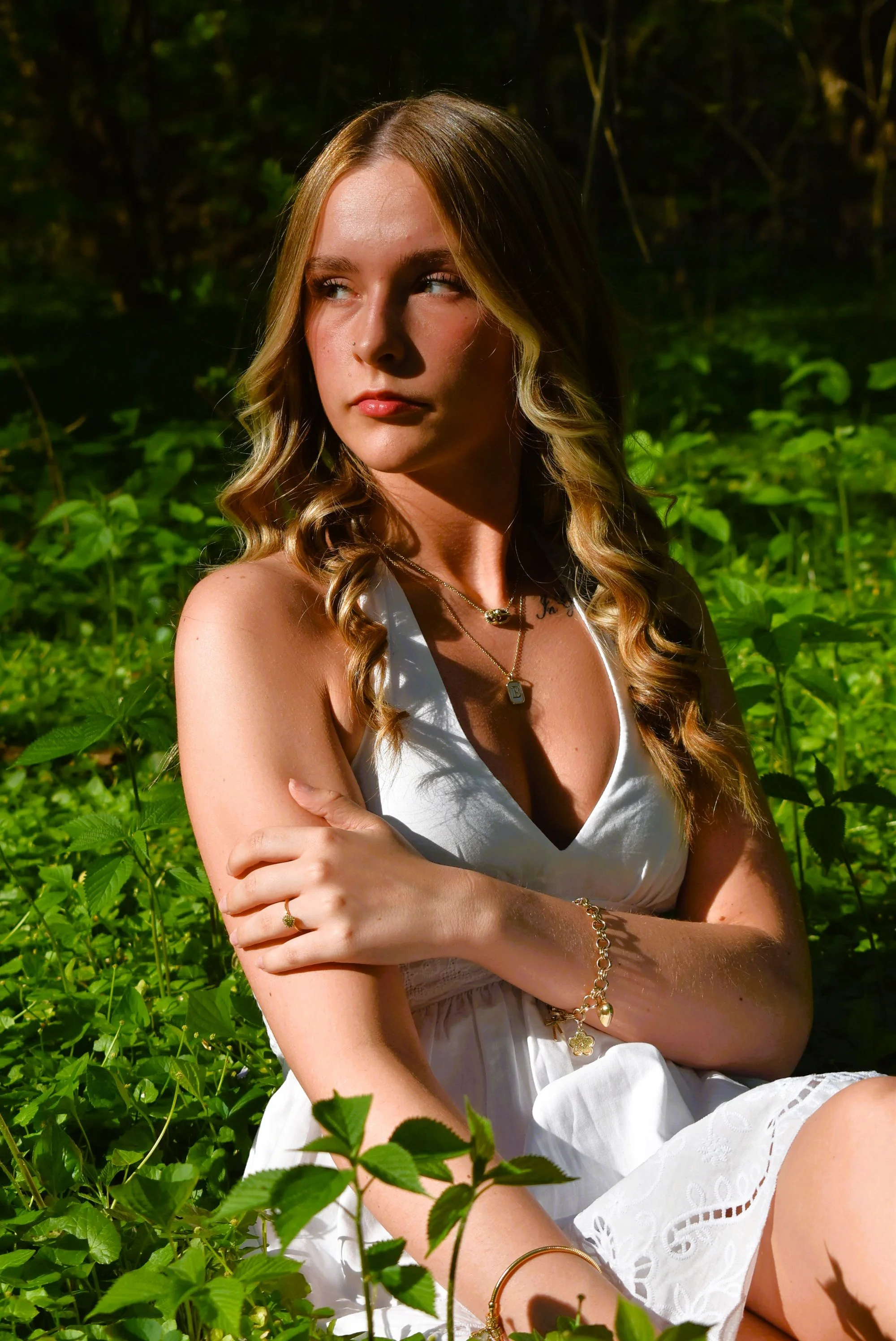 Young woman with wavy blonde hair sitting among green plants, wearing a white sleeveless dress, gold jewelry, and looking to the side in a natural outdoor setting.