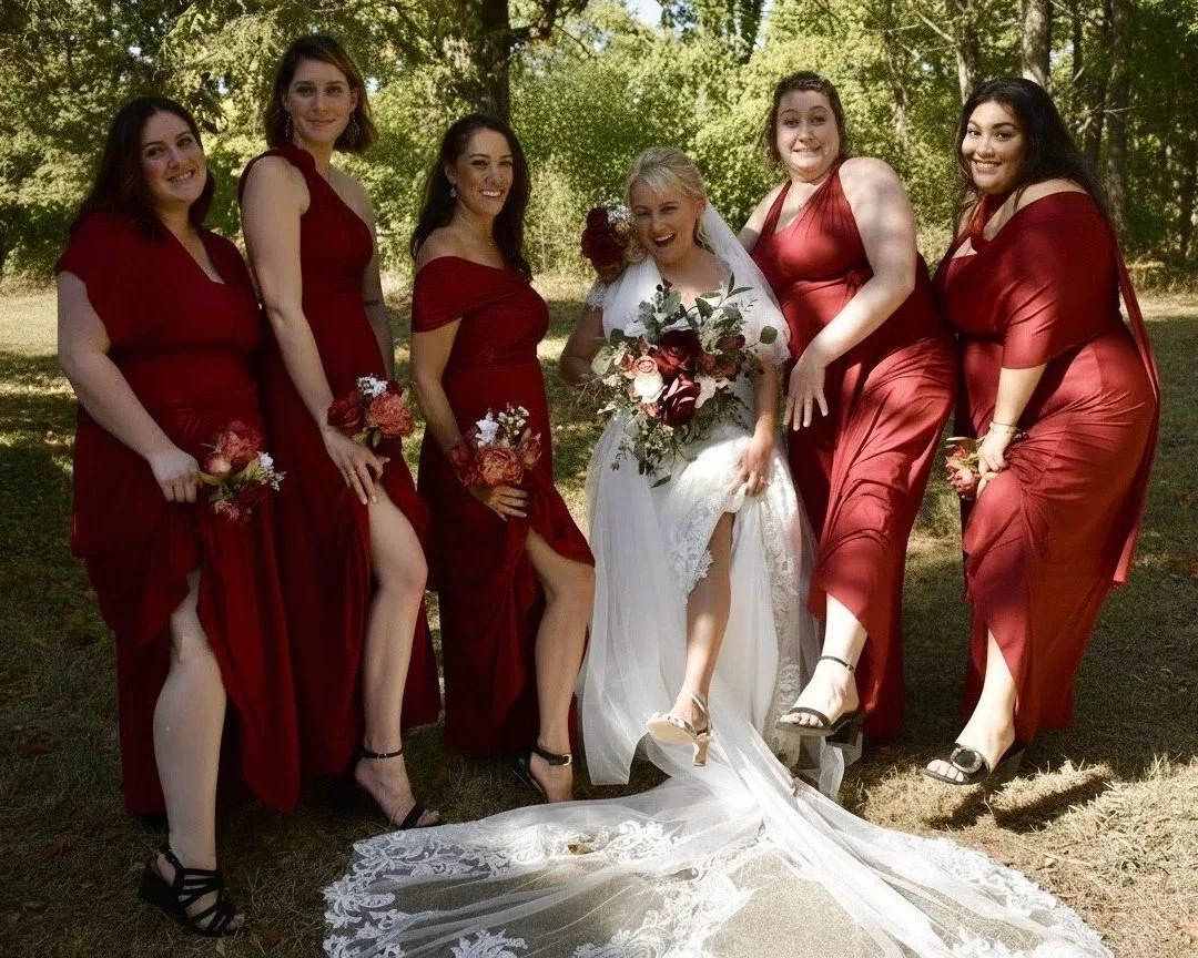 Bride with seven bridesmaids outdoors, all dressed in red gowns, holding floral bouquets, with trees in the background.