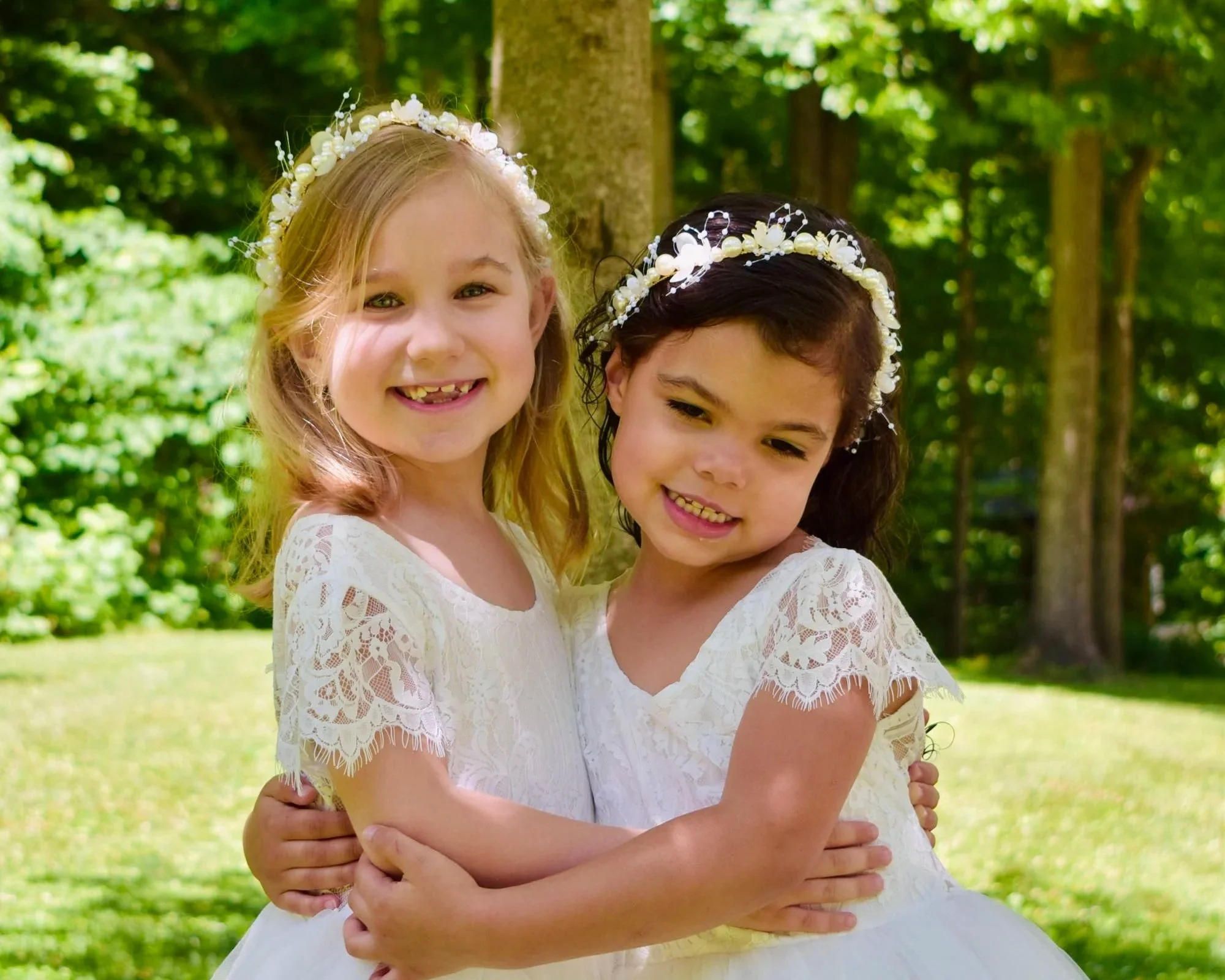 Two young girls wearing white lace dresses and pearl flower crowns hugging outdoors in a green, wooded area.