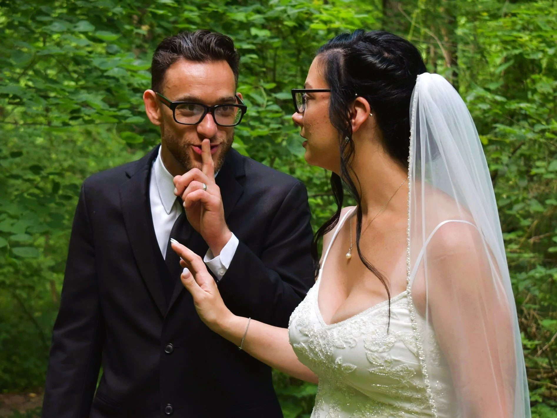 A bride and groom dressed in wedding attire in a wooded outdoor setting. The groom, wearing glasses and a black suit, is holding a finger to his lips in a shushing gesture, while the bride, also wearing glasses and a white wedding dress with a veil, is gently touching the groom's chest.
