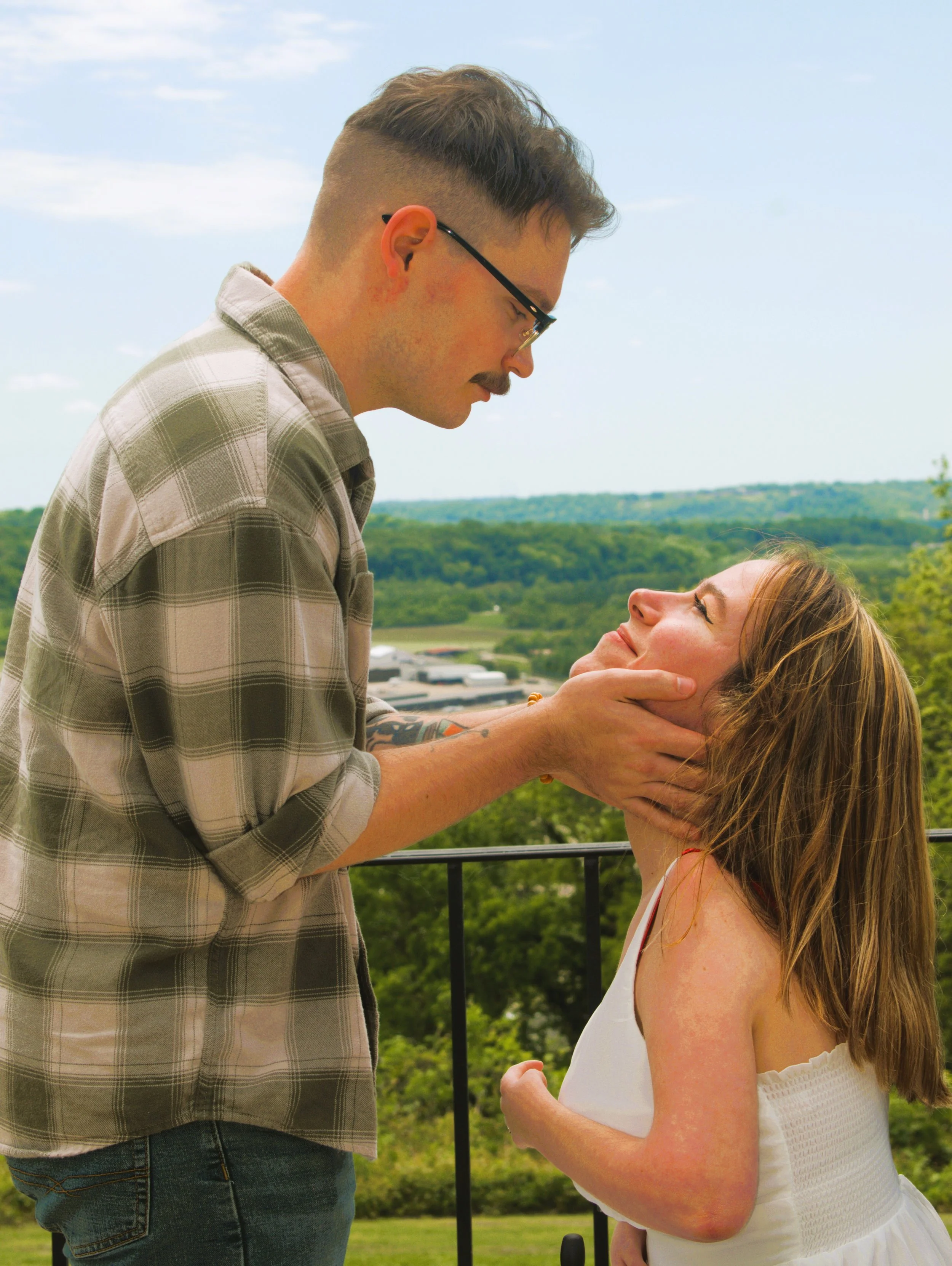 A man with glasses and a plaid shirt holding a woman's chin, looking into her eyes against an outdoor background with greenery and a cloudy sky.