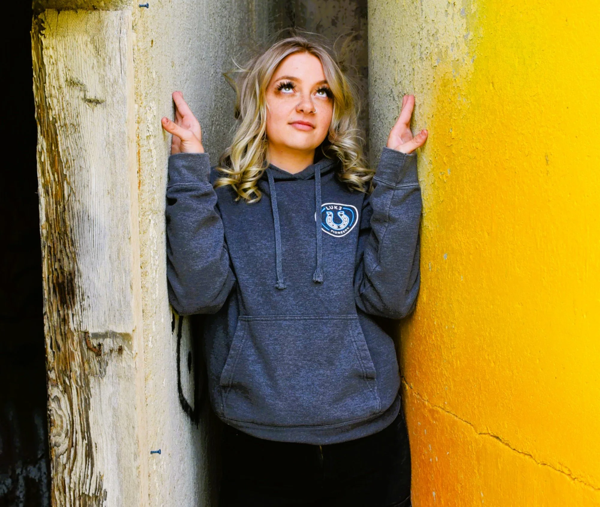A young woman with blonde, curly hair standing between two walls, one wooden and worn, the other painted bright yellow. She is wearing a dark gray hoodie with a circular emblem on it and has a neutral expression, looking upward with her eyes and hands resting against the walls.