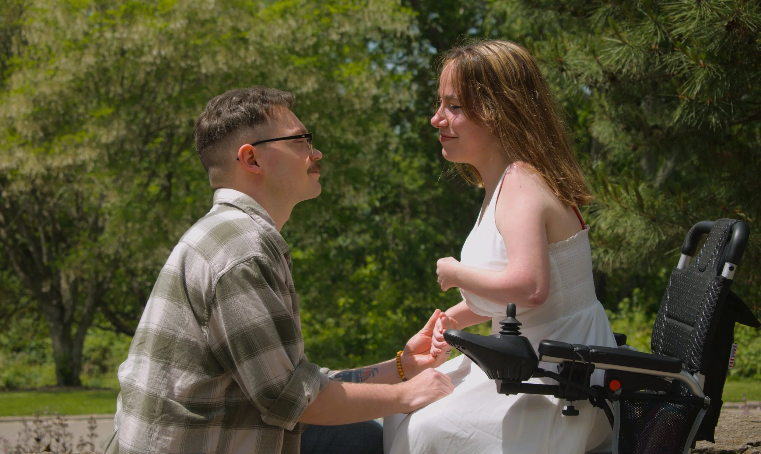 A man with glasses kneeling and holding hands of a woman with a disability in a wheelchair, looking into each other's eyes outdoors surrounded by trees.