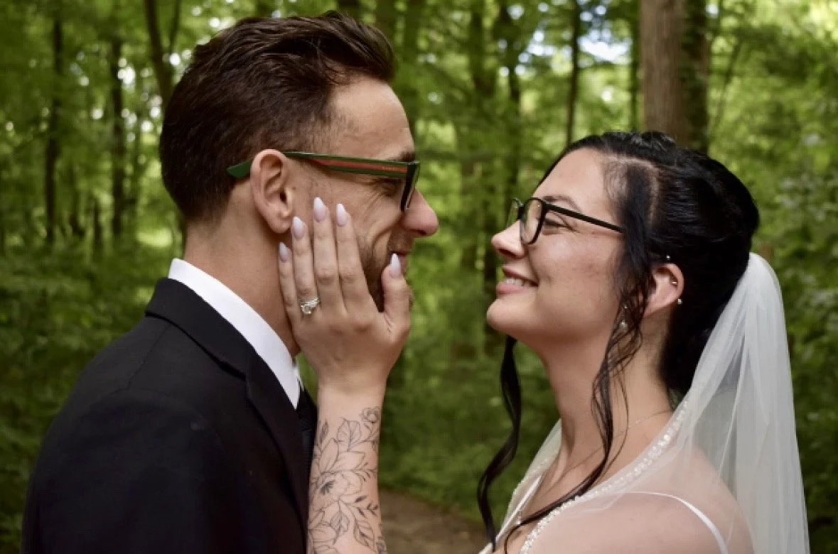 A bride and groom in wedding attire sharing a joyful moment in a forest, with the bride holding the groom's face.