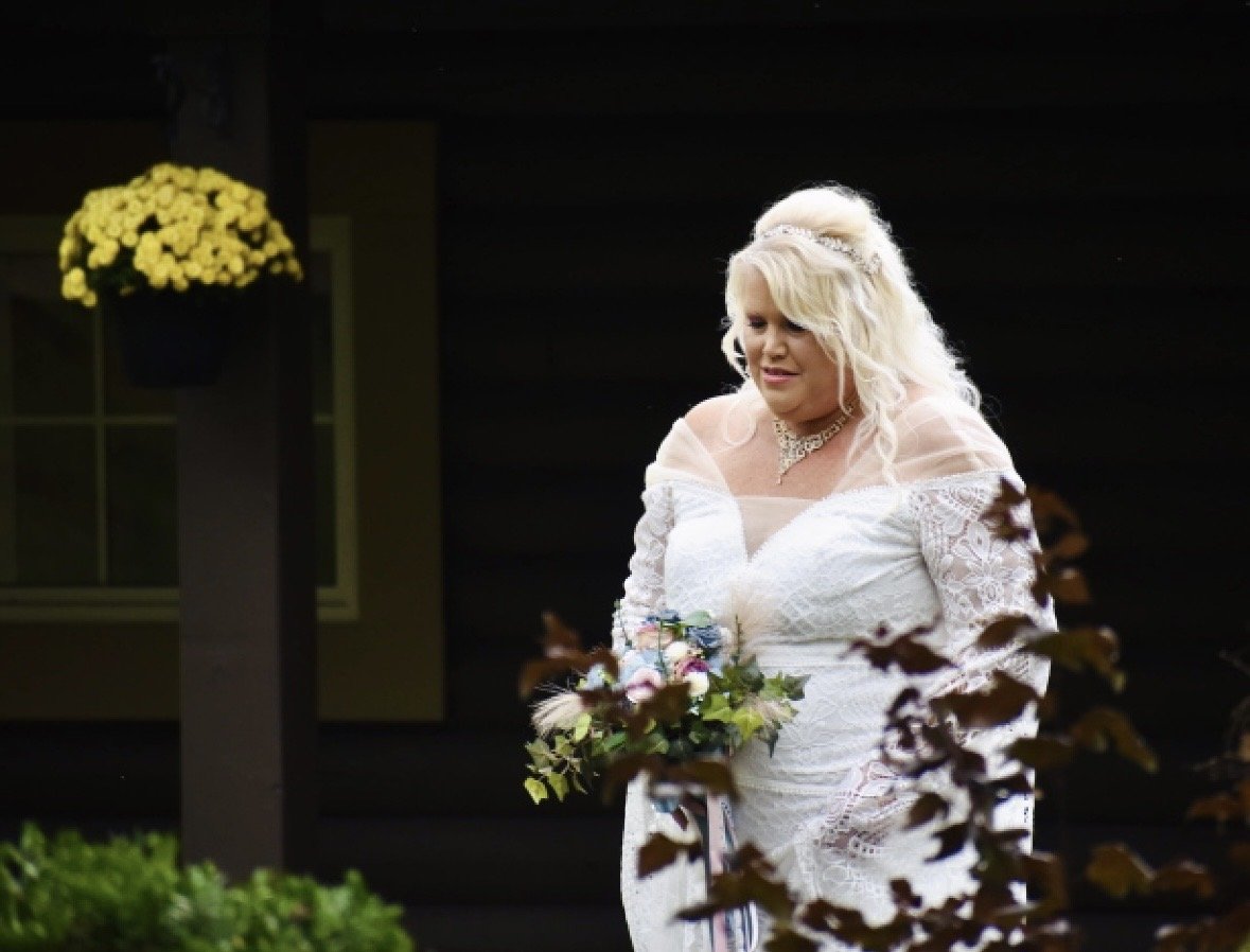 A woman in a white lace dress holding a bouquet of flowers, standing outside near a dark cabin with a yellow potted plant on the wall.