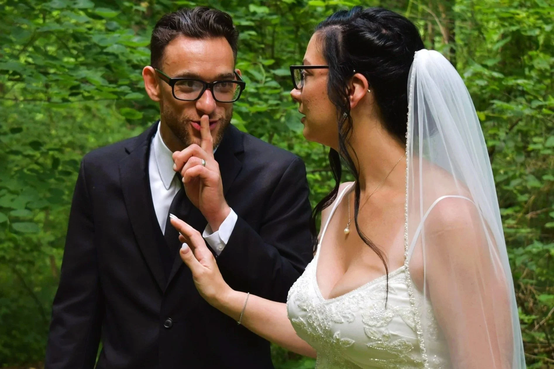 A couple on their wedding day in a lush green outdoor setting, with the groom whispering a secret and the bride smiling, both wearing glasses and wedding attire.
