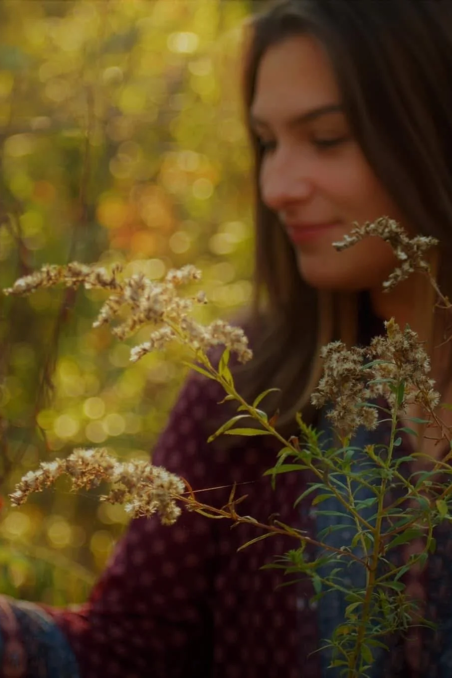 A woman with long brown hair smiling while holding a plant with small white flowers, outdoors during fall with blurred trees and sunlight in the background.