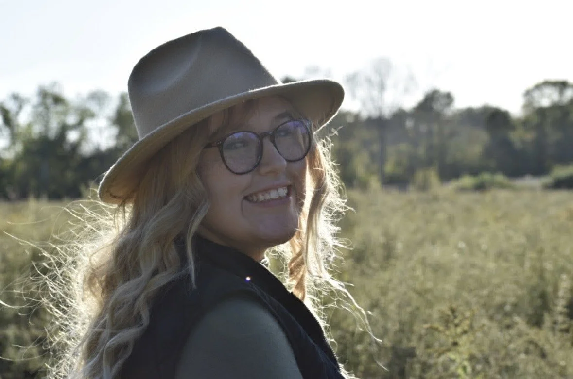 A woman with blonde hair, glasses, and a wide-brimmed hat smiling outdoors in a natural setting with trees and plants in the background.