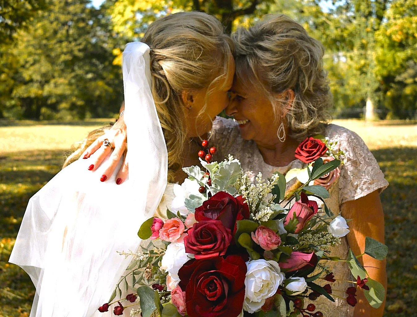 Two women, a bride and an older woman, with foreheads touching and smiling while holding a bouquet of roses and other flowers outdoors in a park with autumn-colored trees.