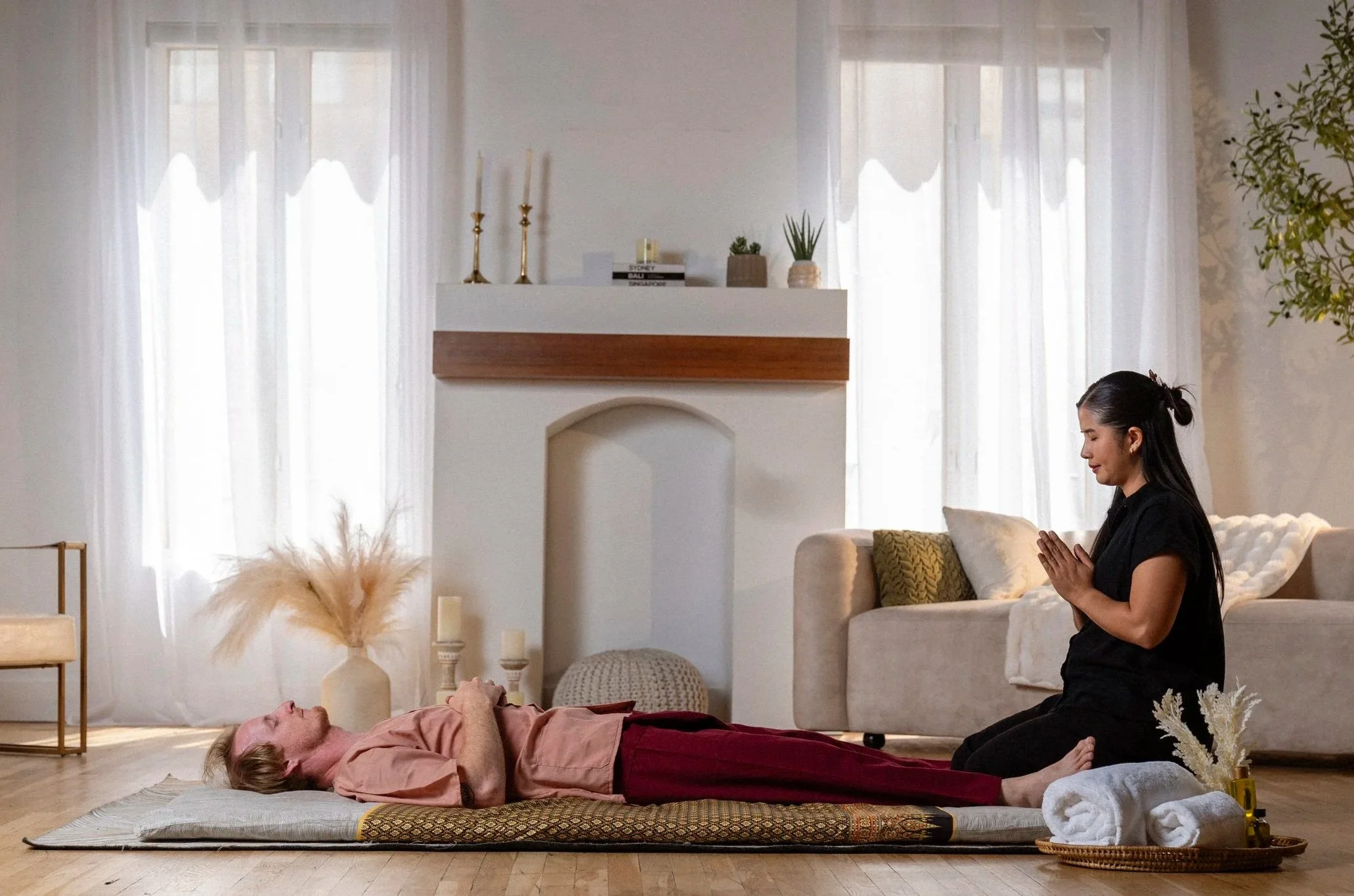 Therapist in prayer pose beside client resting on Thai mat, representing the Sen Flow Body practice.